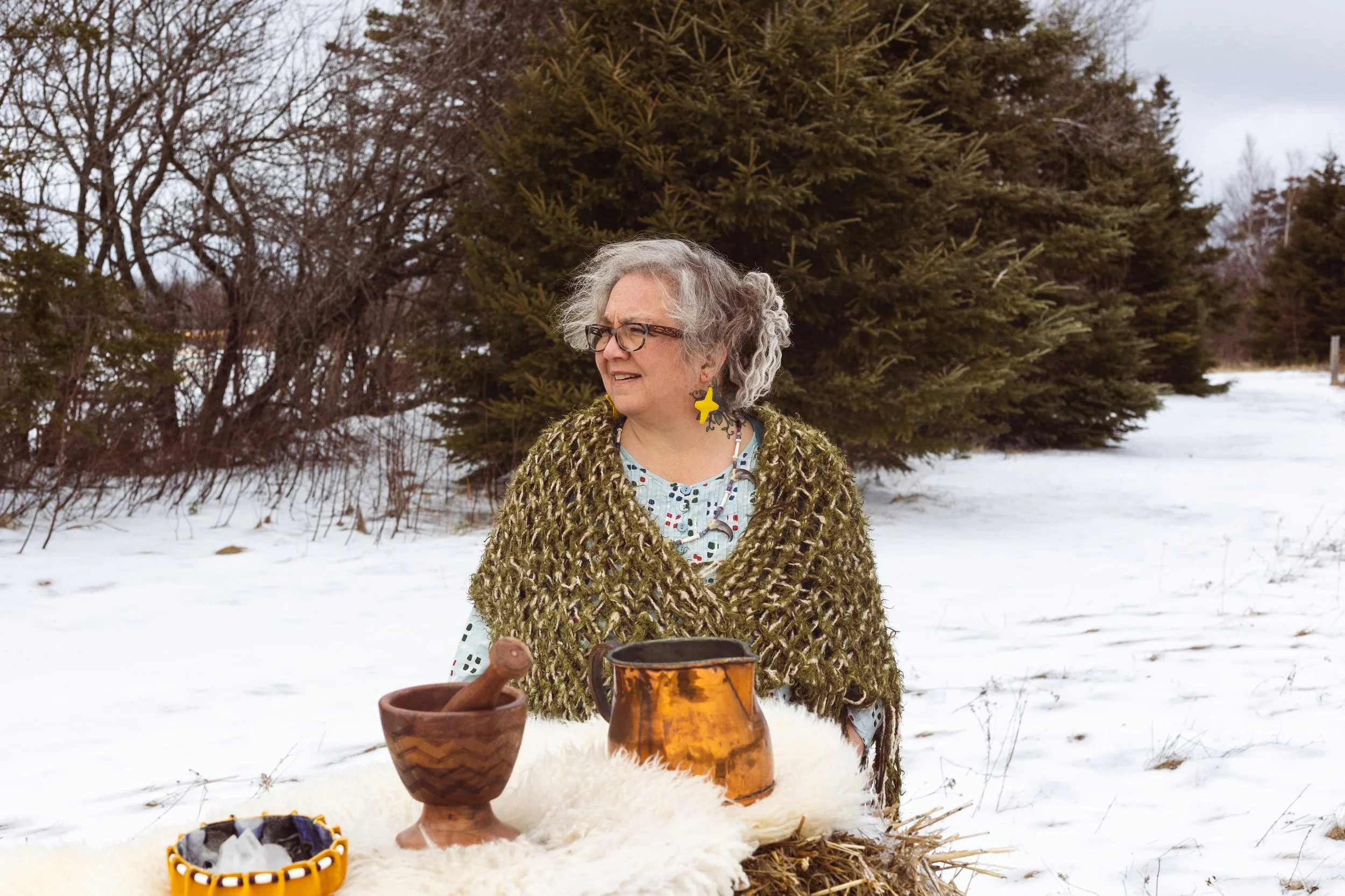 A woman with gray, curly hair wearing glasses, a green textured shawl, and large star-shaped earrings stands outside on snow, with trees in the background. In front of her are three pottery vessels on a white furry cloth, with a small bowl or container filled with various items.