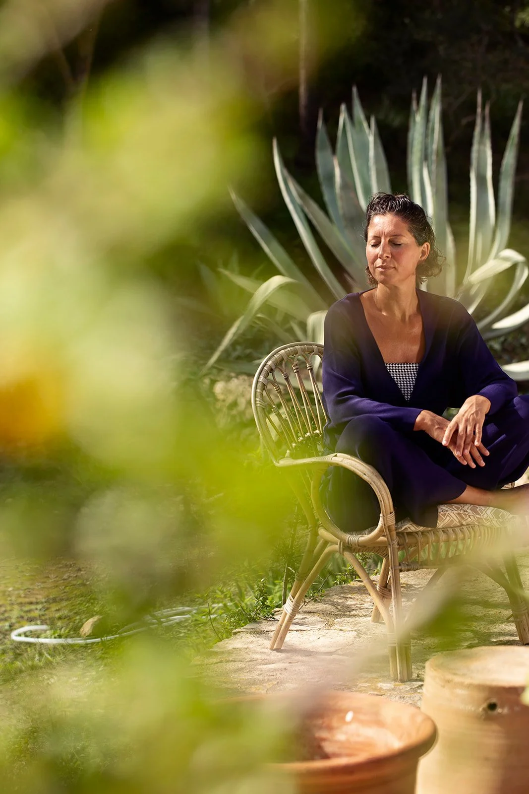 Marianne de Kuyper sitting calmly in a rattan chair in front of a large agave plant, eyes closed, in a Mallorca garden.