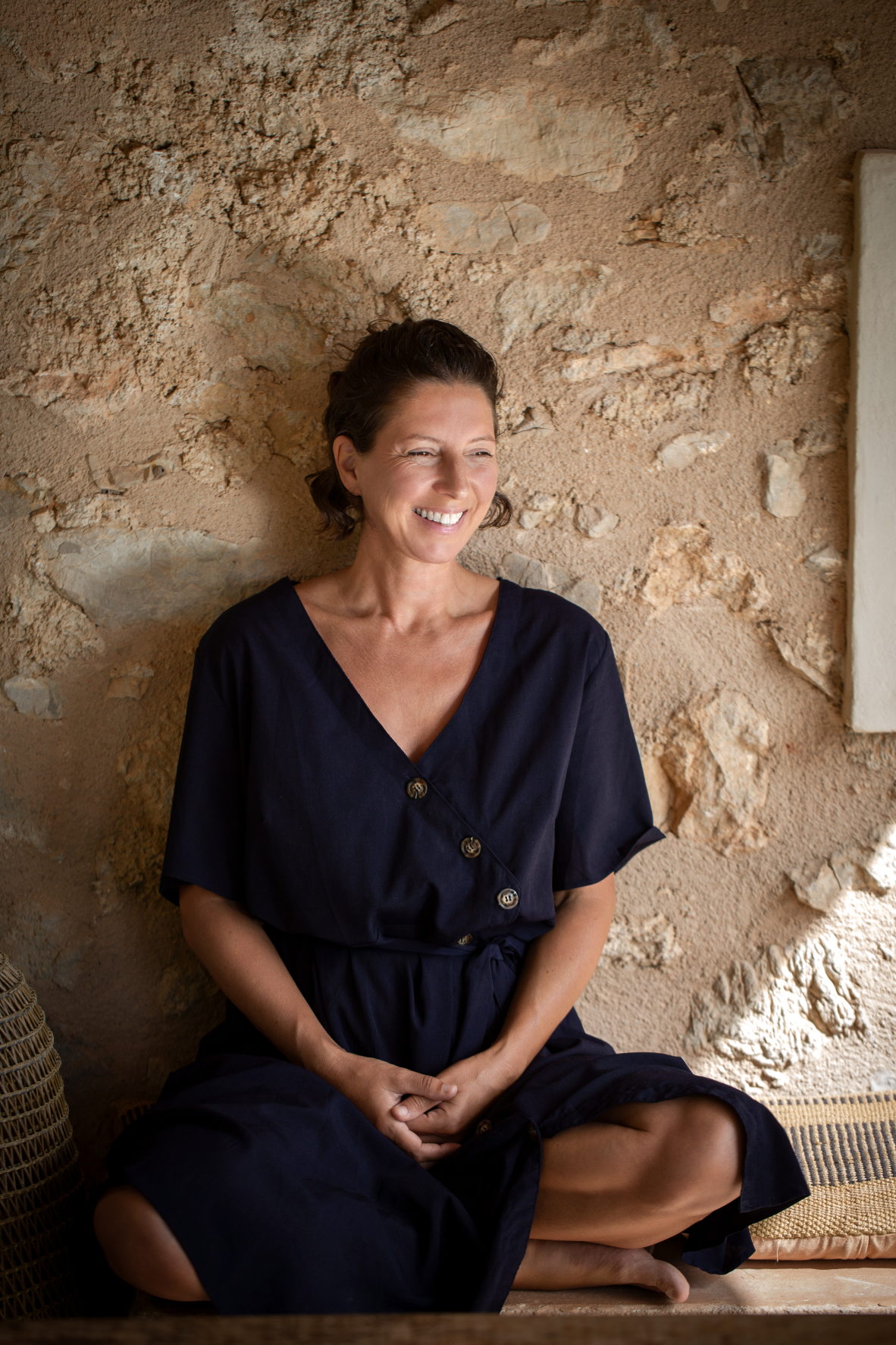 Marianne smiling warmly, sitting barefoot against a stone finca wall in Mallorca