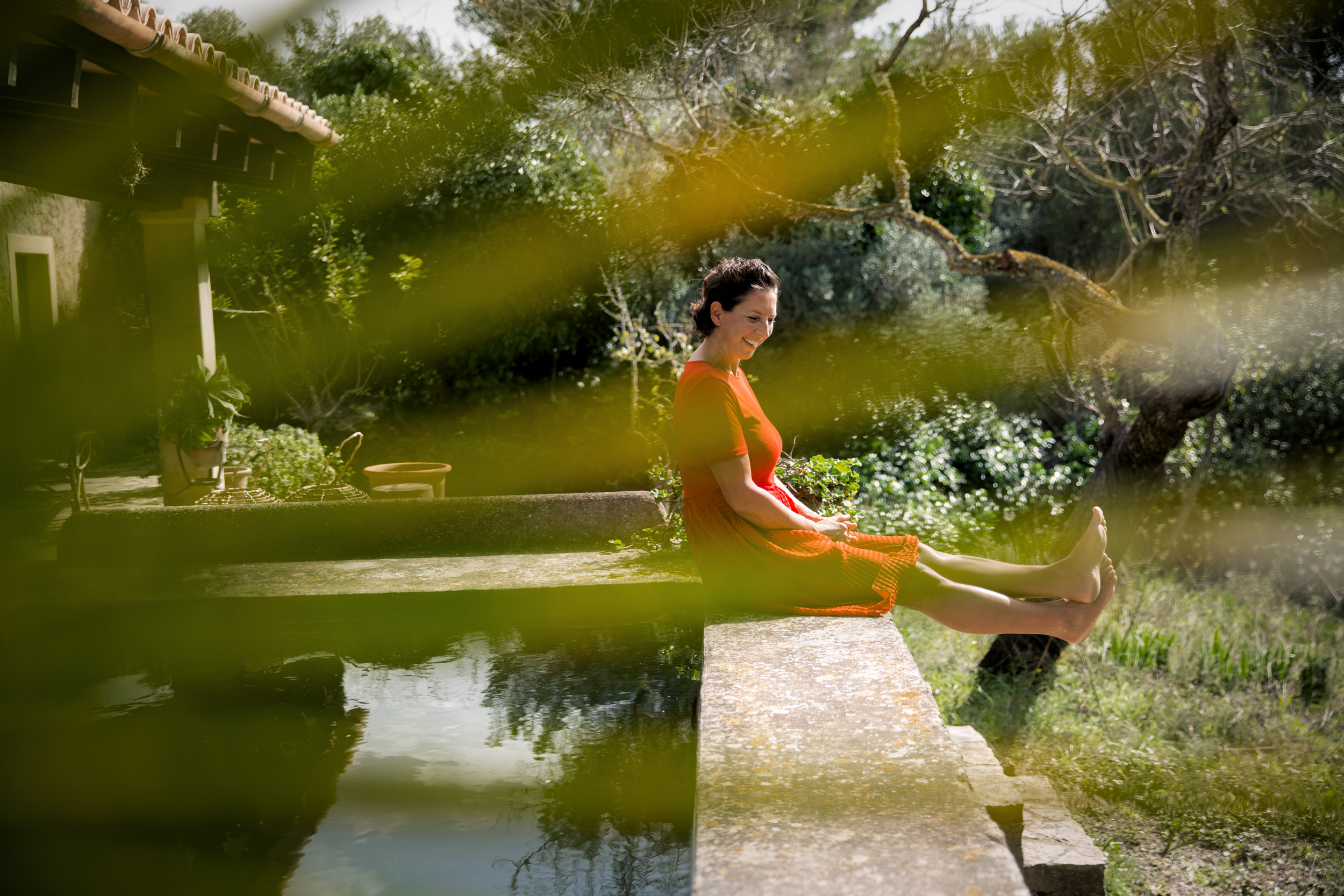 Marianne de Kuyper sitting barefoot beside a pond in a Mallorca garden, surrounded by cacti and succulents.