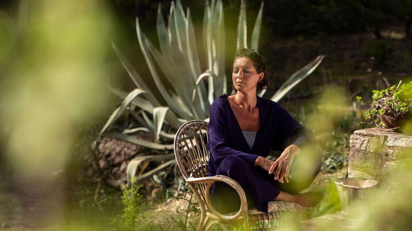 Marianne sitting cross-legged and reflective in a rattan chair among agave plants in Mallorca