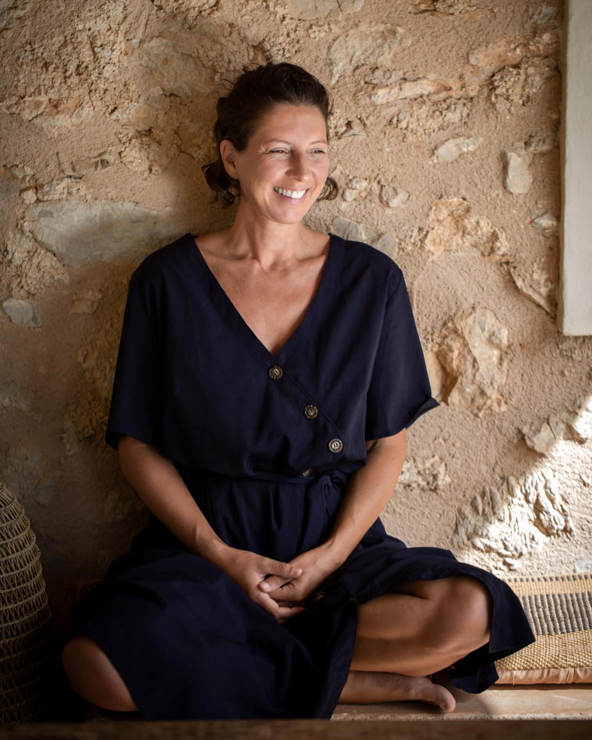 Marianne de Kuyper, coach and yoga teacher at Studio20, sitting cross-legged and smiling against a rustic stone wall on Mallorca.