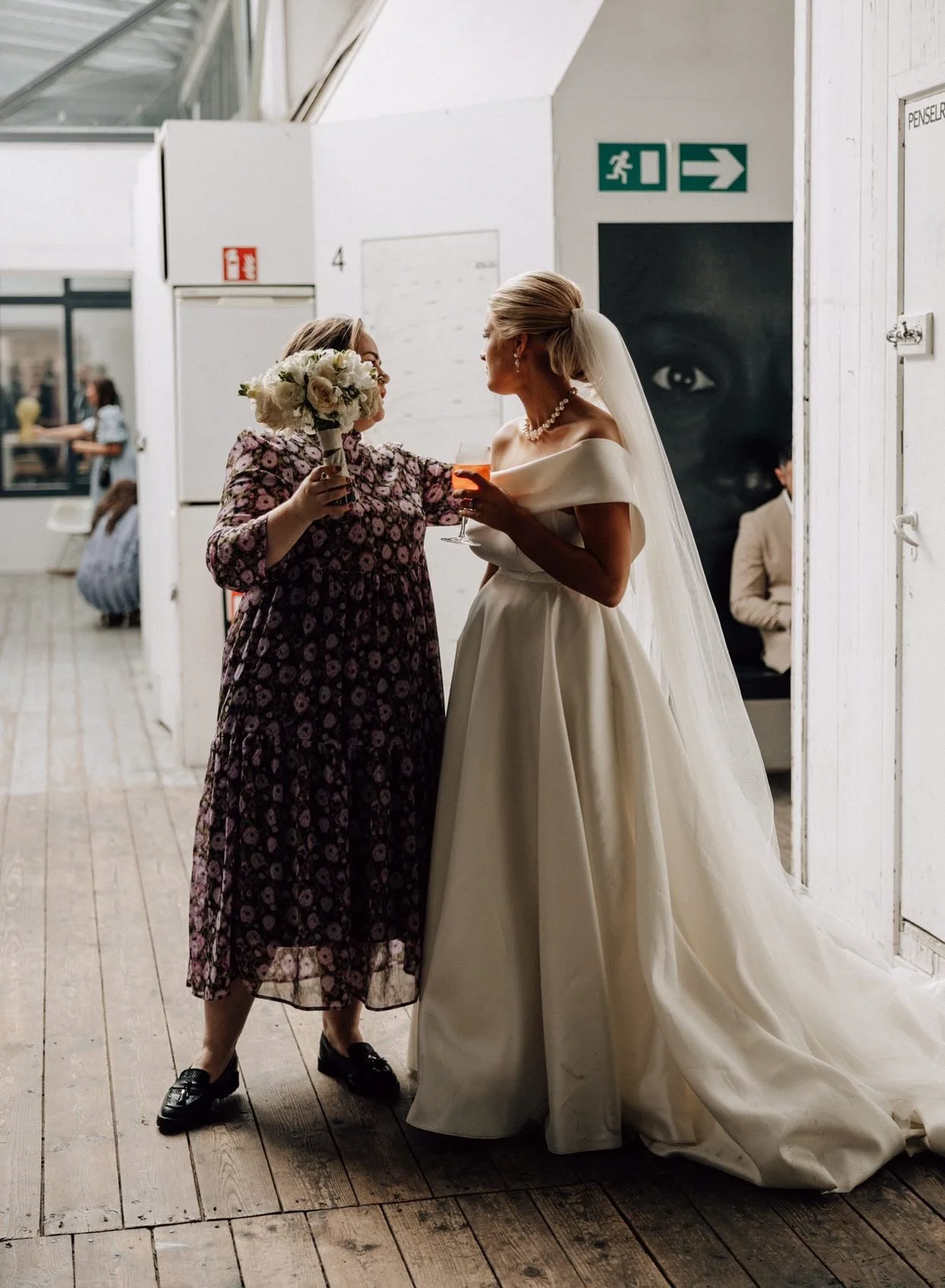 Bryllupsplanlægger Cat Bjørnholt hjælper brud på bryllupsdagen. De skåler sammen indendørs, hvor bruden bærer hvid kjole og blomsterbuket.