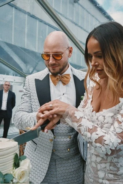 Et par i festkjoler skærer en bryllupskage ved et bord, med andre gæster i baggrunden.