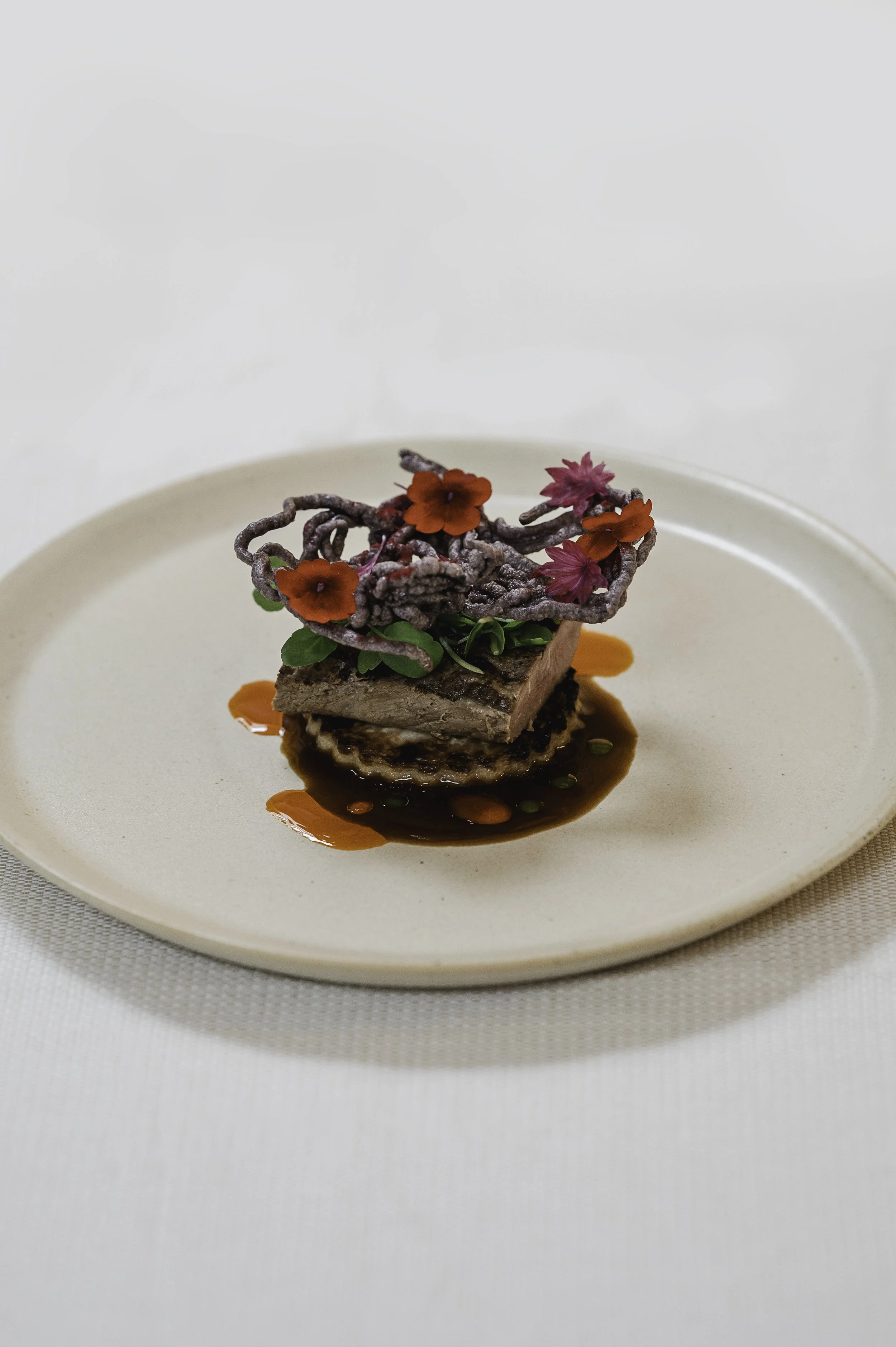 Eksklusiv anretning med kød, sauce, grønt og spiselige blomster på en beige tallerken.
