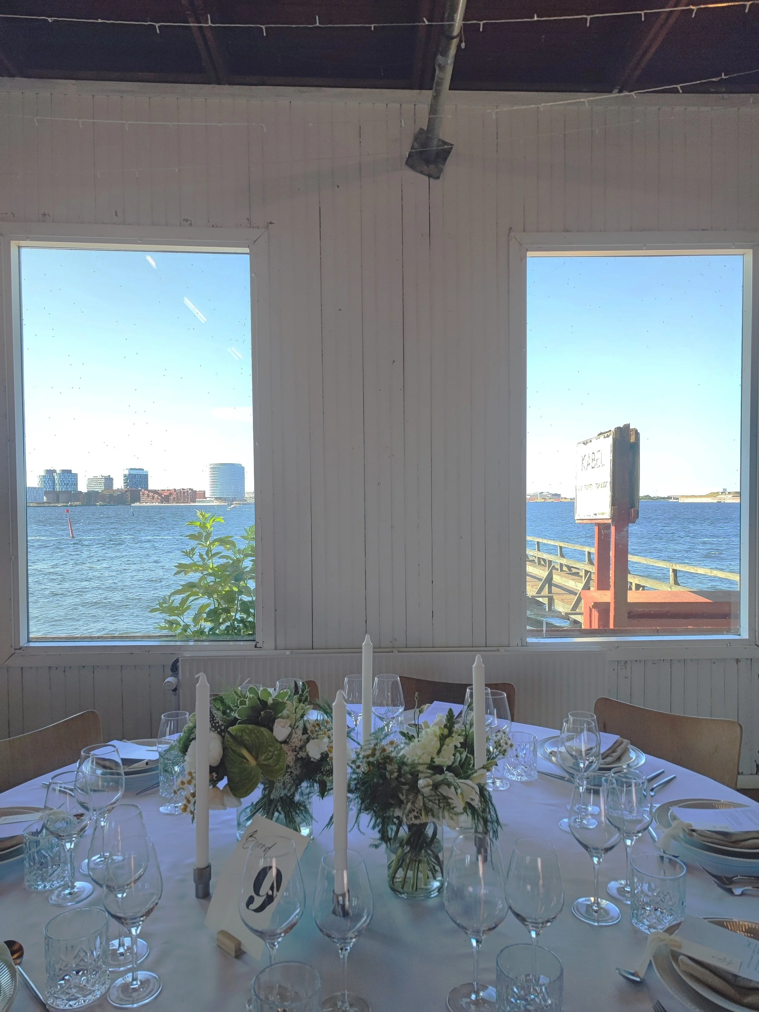 Indretning med bord dækket til fest, blomsterdekorationer og stearinlys, med udsigt over vandet gennem to store vinduer