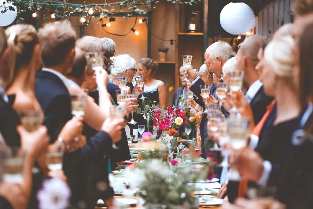 Gæster til fest eller bryllup til at skåle med drinks ved et langt serveret bord med blomsterdekorationer, i en indendørs eller udendørs festlig setting.
