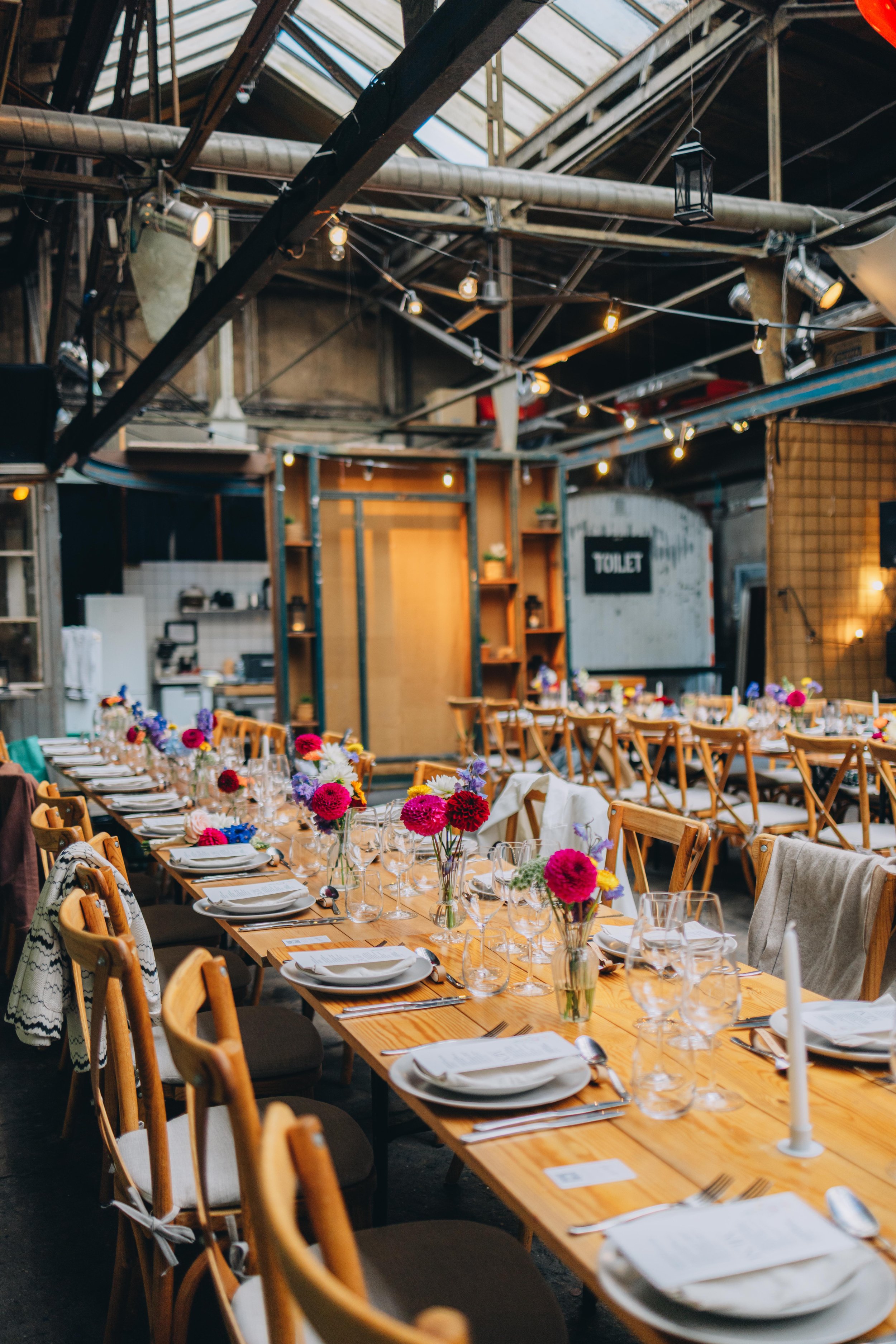 Bliss CPH – Festbord med blomsterdekorationer i et industrielt lokale med træstole og dækket med tallerkener, glas og bestik