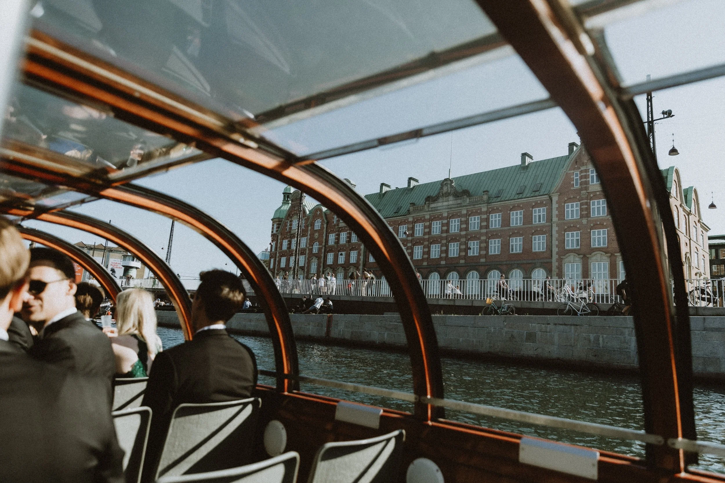 Mennesker på en bådtur langs en kanal med historiske bygninger i baggrunden, set fra bådens indre glasoverdækkede område.