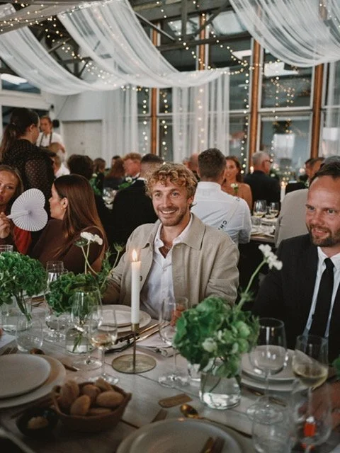 Mennesker til en festmiddag, dekoreret med blomster og stearinlys, indendørs, med hvide gardiner og lys i loftet.