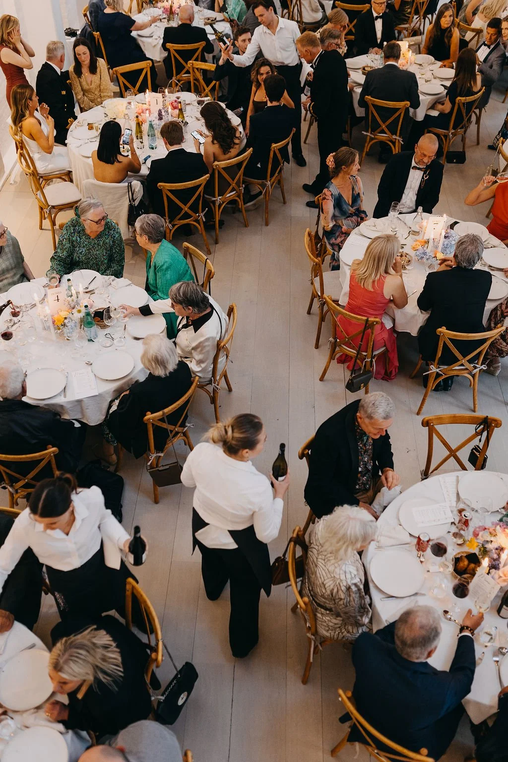 Billede af en elegant festmiddag med mange gæster, der sidder ved rundt bord dækket med hvide duge, blomsterdekorationer og serveringsservice. Gæsterne er klædt formelt, og nogle personer serverer mad og drikke.
