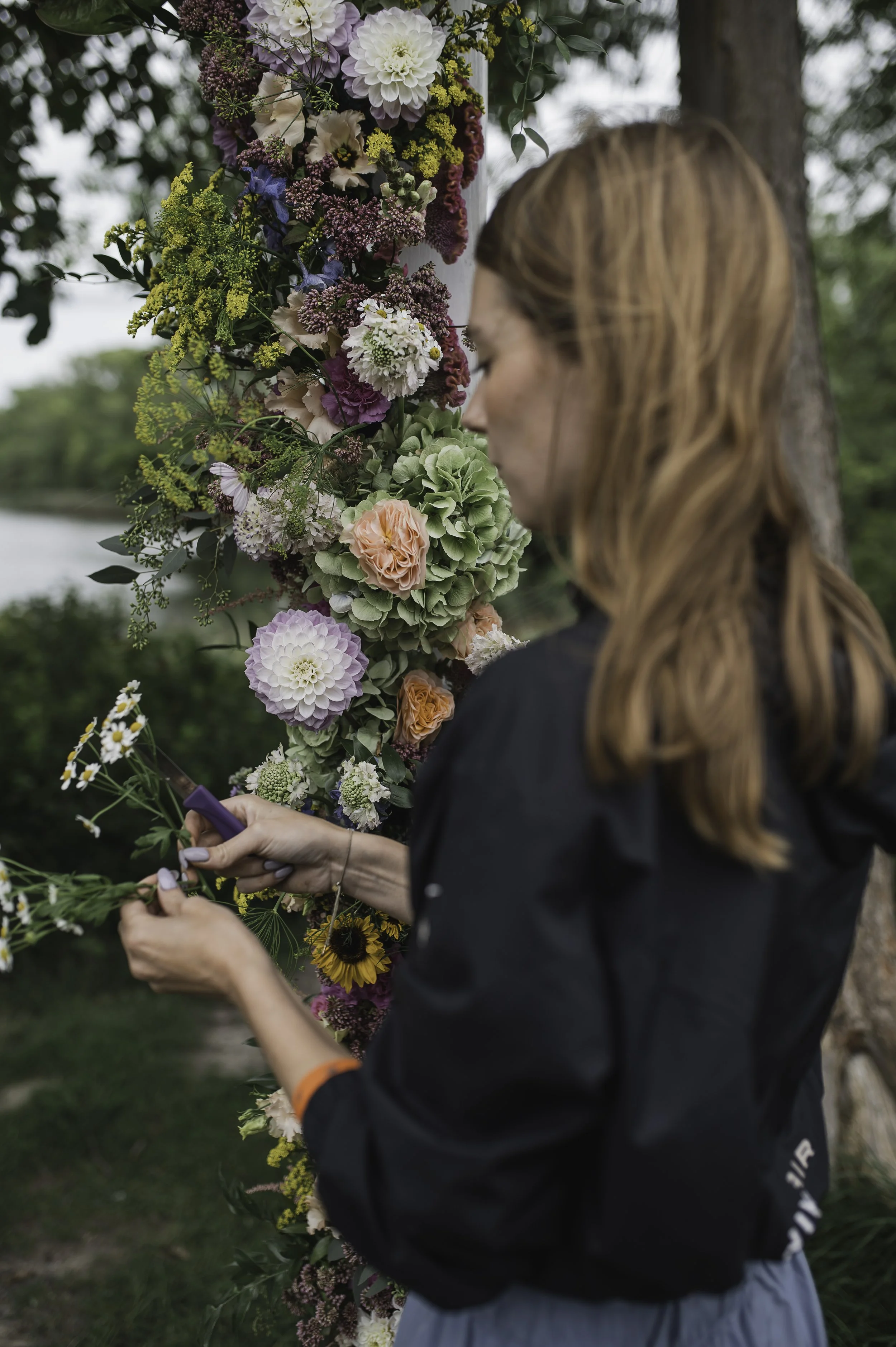 Kvinde med brunt hår arbejder med blomster foran en blomsteropsats udendørs.