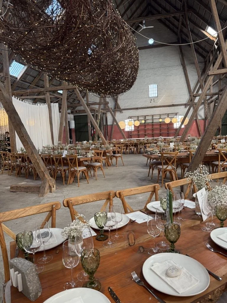 Indretning til fest i en stor, rustik lade med træbjælker, rund og firkantet borde dækket til middag, blomsterdekorationer, store césteterlygter, store papballoner, og et stort kunstigt rede ophængt i loftet med små lys.