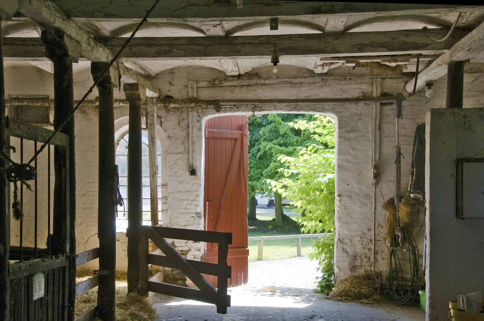 Bliss Cph - Indendørs del af Søllestedgård med åbent rødt dør, grønne træer udenfor, træstolper, hestebokse, og lidt udstyr på væggen.