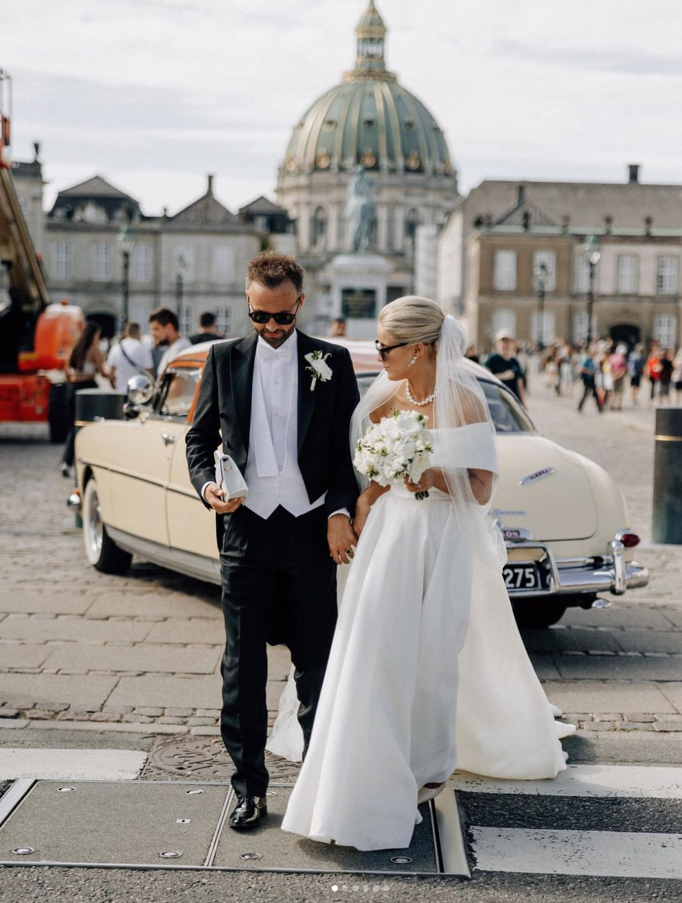 En brud og brudgom går hånd i hånd på gaden, med en vintage bil i baggrunden. Bruden bærer en hvid kjole og holder en blomsterbuket, mens brudgommen har smoking og solbriller. Der er en stor kuppelbygning i baggrunden, og der er flere mennesker i baggrunden.