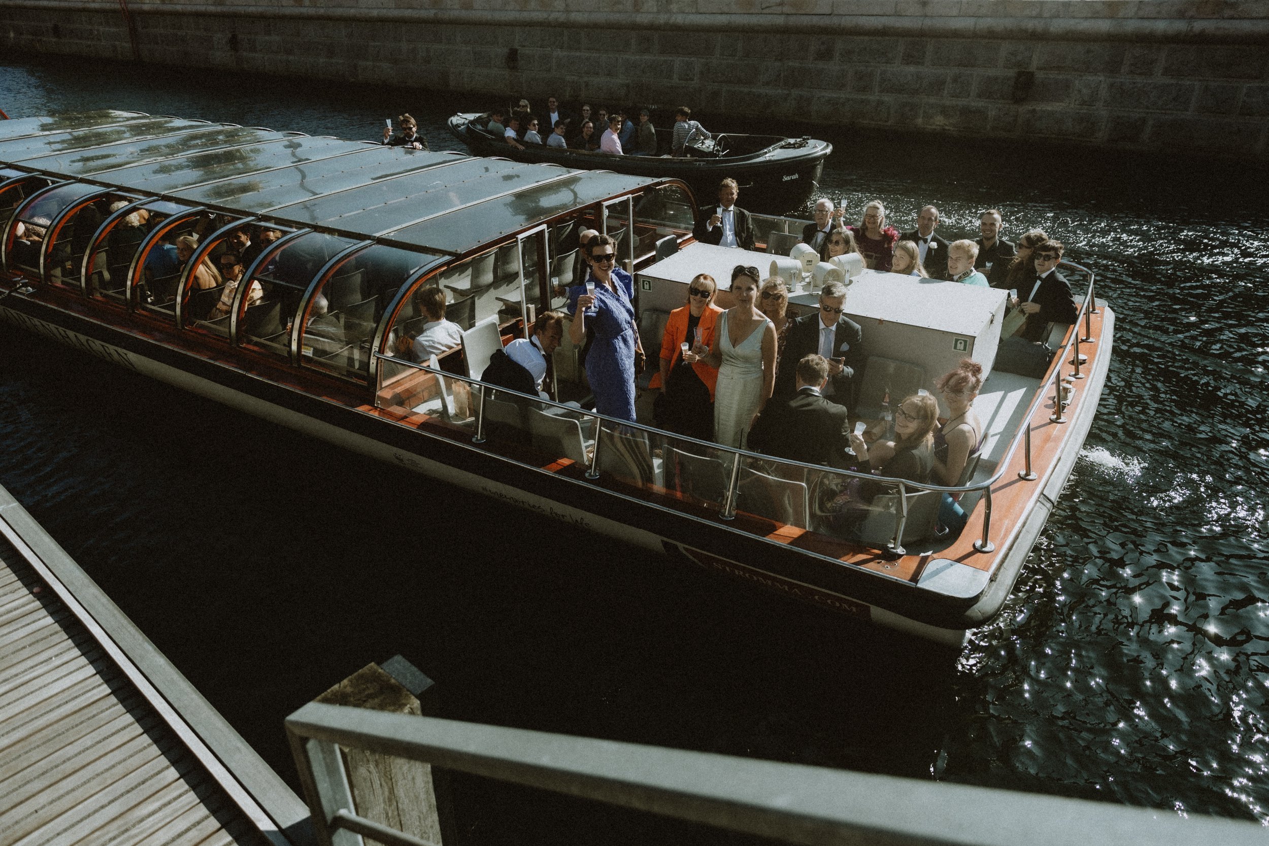 Bliss CPH – En bryllupsgruppe i Københavns havn i fint tøj og en kvinde i brudekjole, ved Kanalrundfart med et glastag