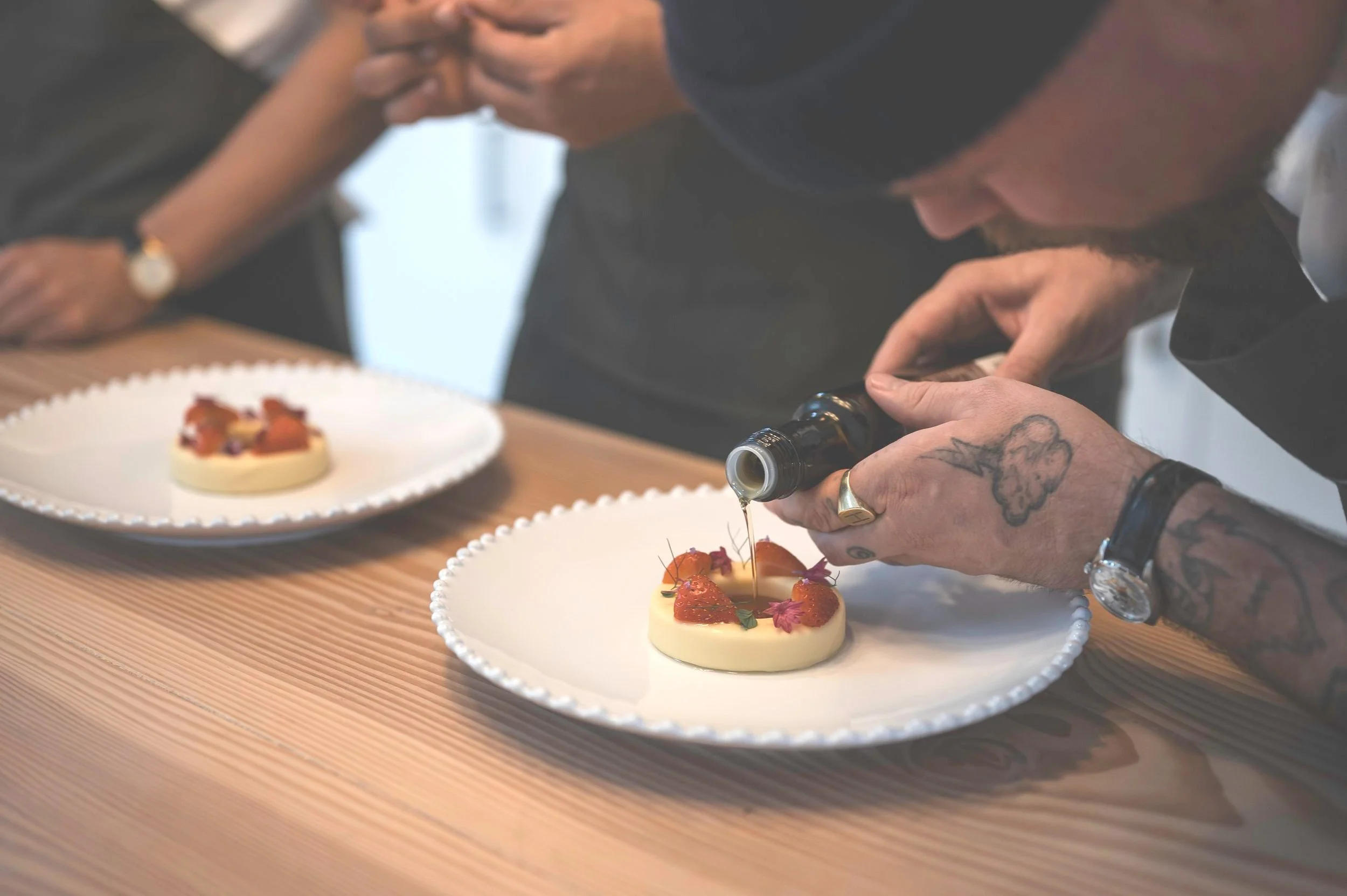 En person med tatoveringer på hånden er ved at dekorere en dessert med sauce, serveret på en hvid tallerken. En anden tallerken med en lignende dessert kan ses i baggrunden.
