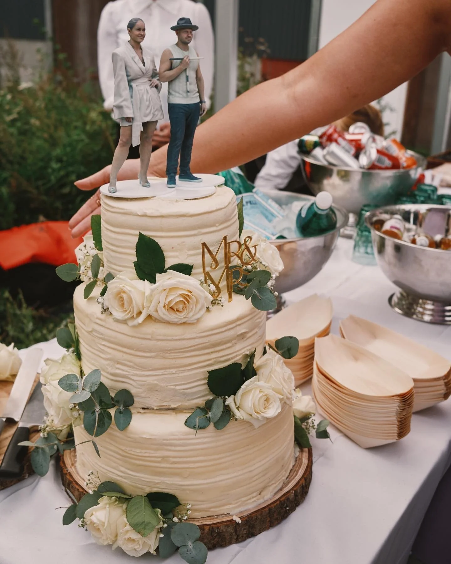 En tre-lags bryllupskage med blomsterdekorationer og en 'M&R' æske som kage-topper med miniaturefigurer af et brudepar på toppen.