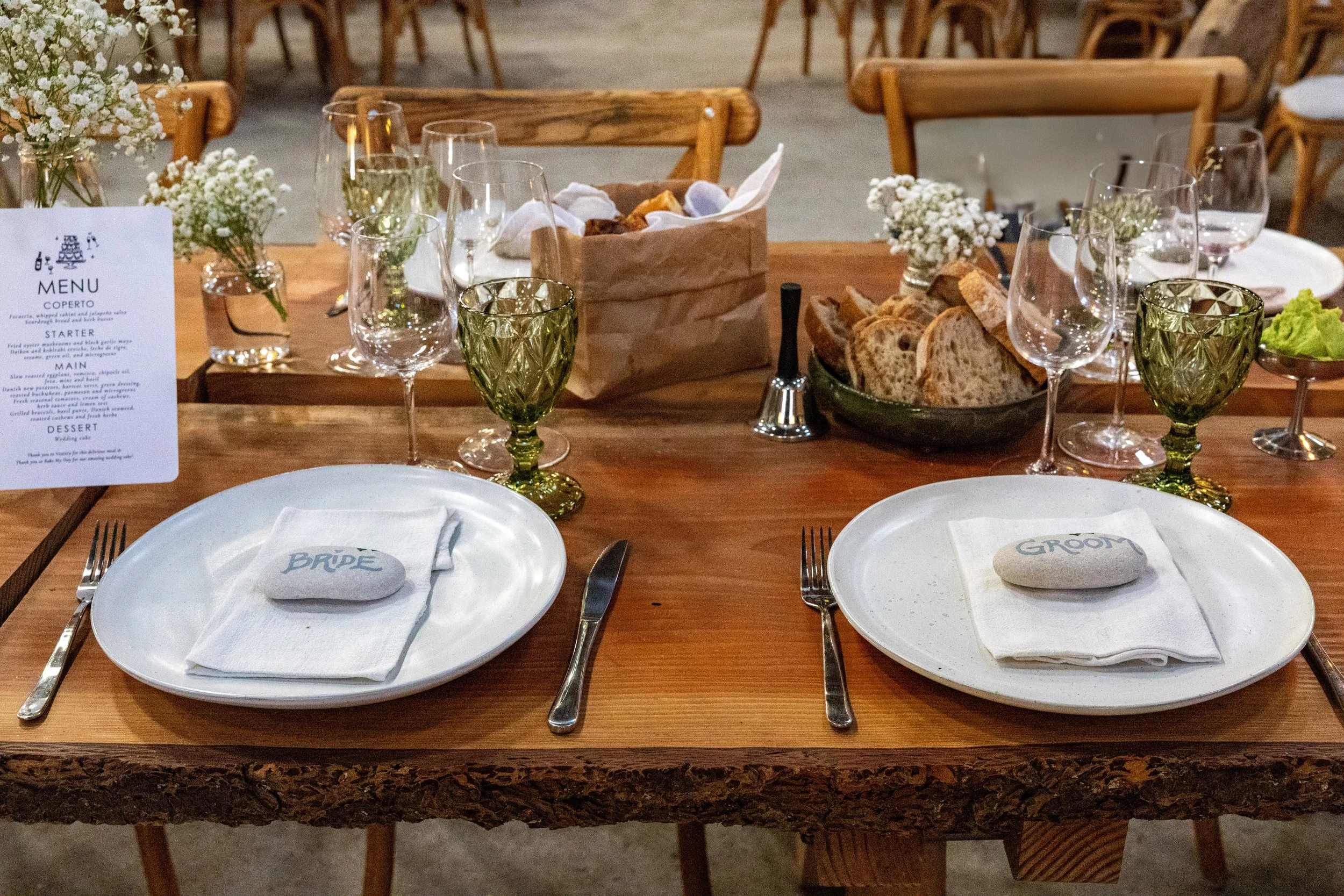 Dækket bryllupsbord med to tallerkener med 'bride' og 'groom' skrevet på sten, bestik, vinglas, blomsterdekorationer, brød og menu kort.