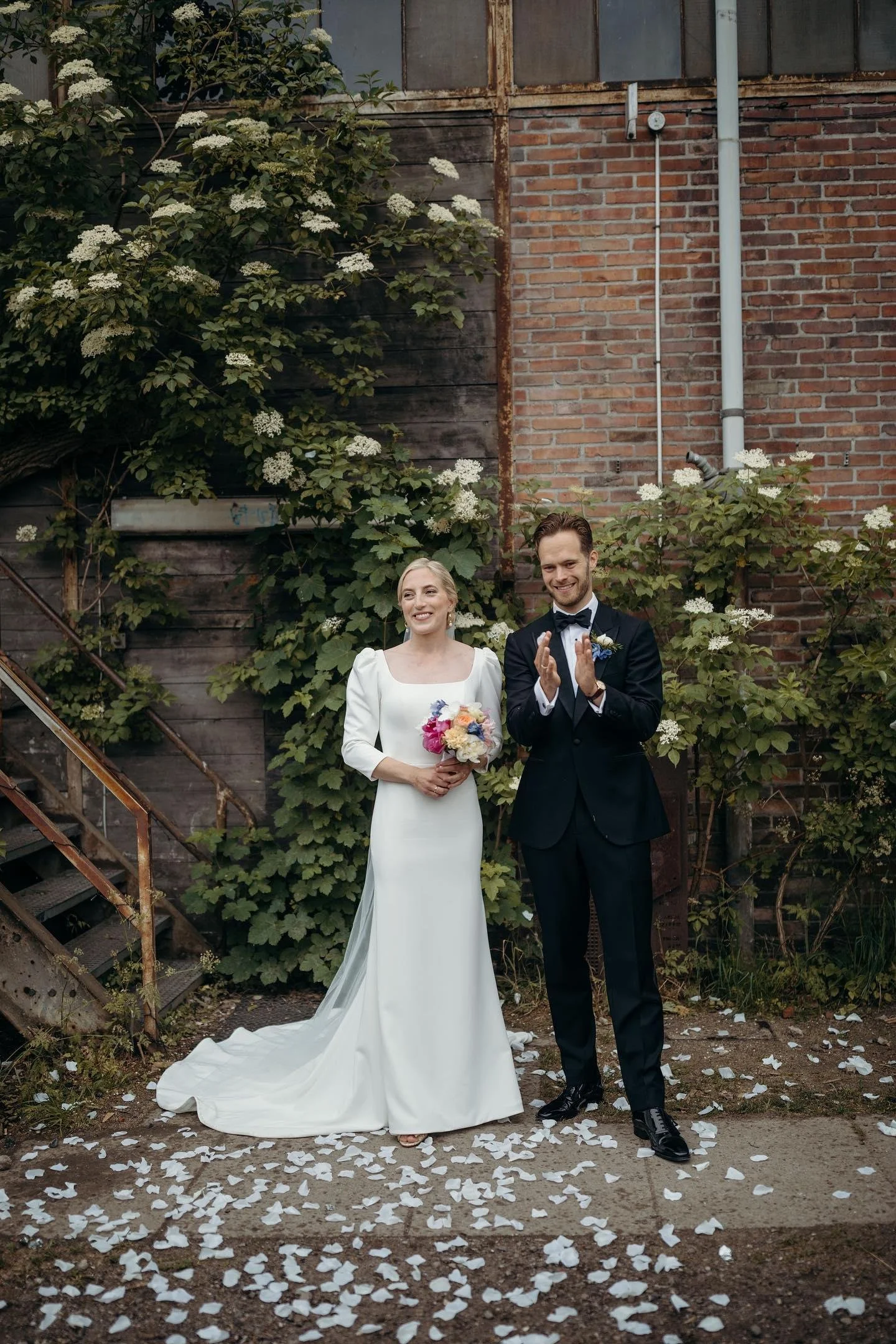 Et brudepar står udenfor foran en mur med grønne planter og hvide blomster. Bruden har en hvid kjole og holder en blomsterbuket, mens gommen bruger en sort jakkesæt og klapper.