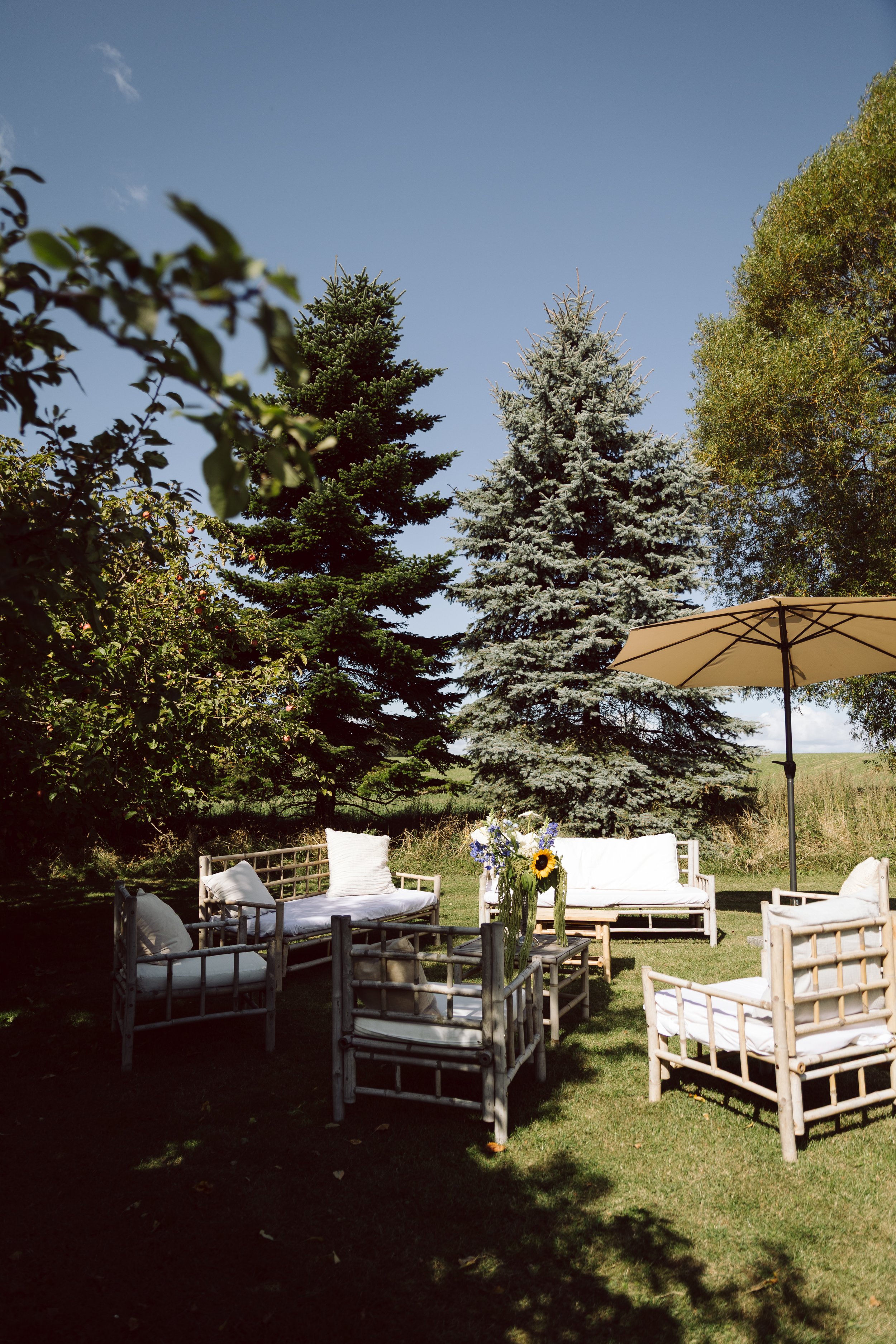 Udendørs havemøbler med hvide puder under en stor parasol, omgivet af grønne træer og en klar blå himmel.
