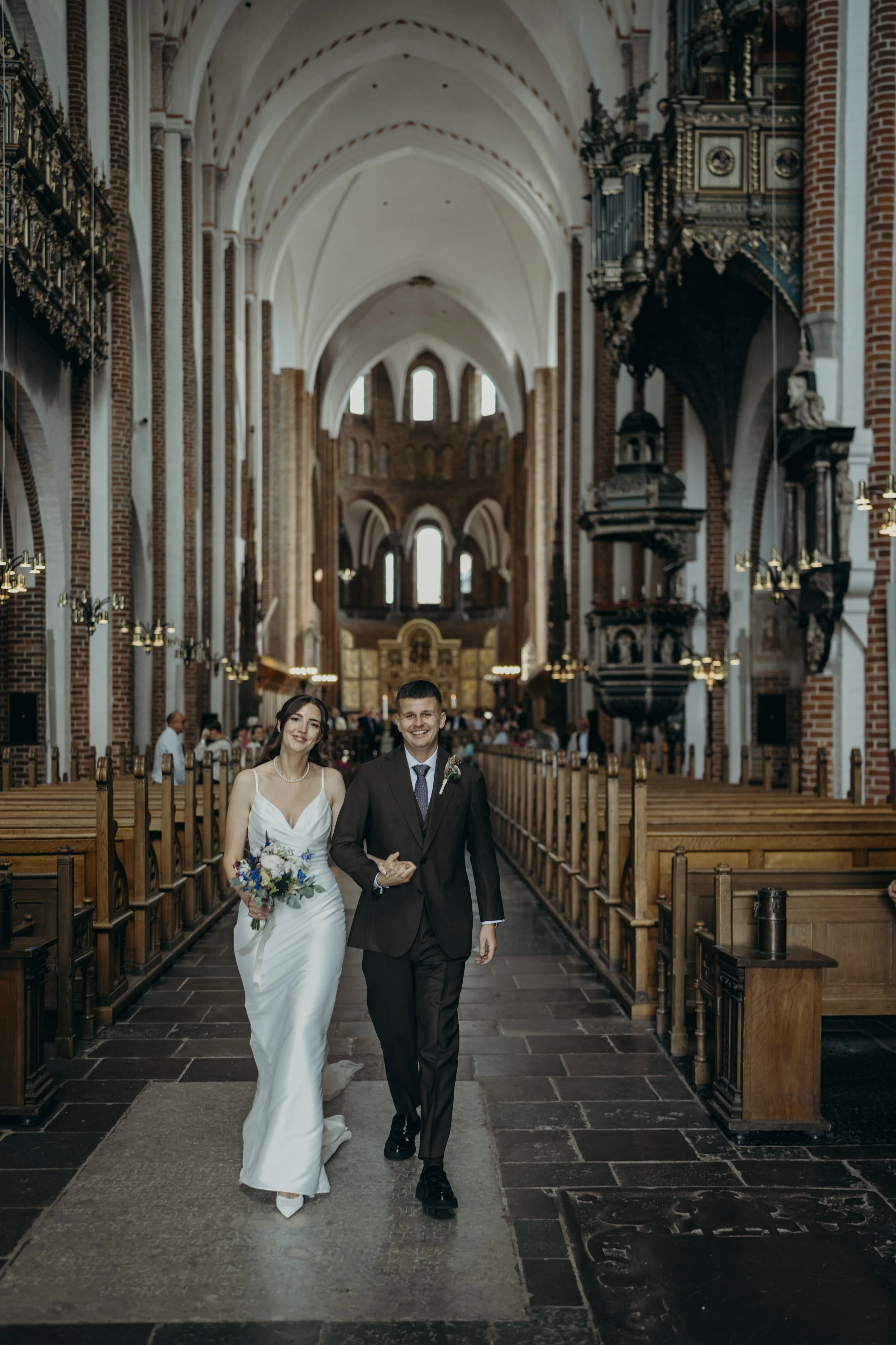Kærligt par i kirke, den ene i hvid brudekjole med blomsterbuket, den anden i sort jakkesæt, går ned ad midtergangen.