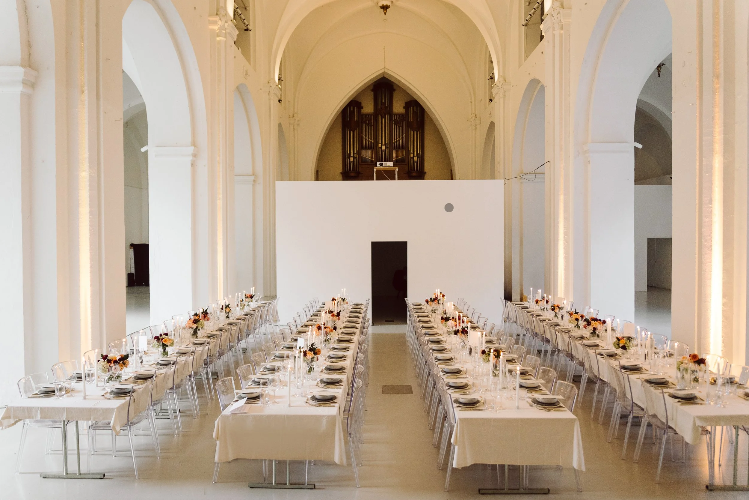 Bryllupsopsætning på Nikolaj Kunsthal med gennemsigtige stole, fire lange borde dækket med hvide duge, stearinlys og KHWürtz. Kontrasttallerkener, glas og blomsterdekorationer i en stor, arkitektonisk kirke med hvide vægge og høje, buede lofter.