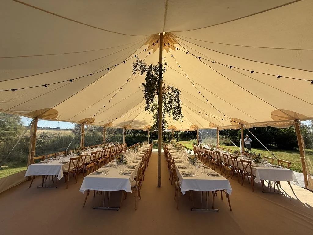Et luksustelt dekoreret af eventplanlæggeren med lange borde dækket med hvide duge og smukke blomsterdekorationer, åbne sider med udsigt til lysegrønne marker og natur.