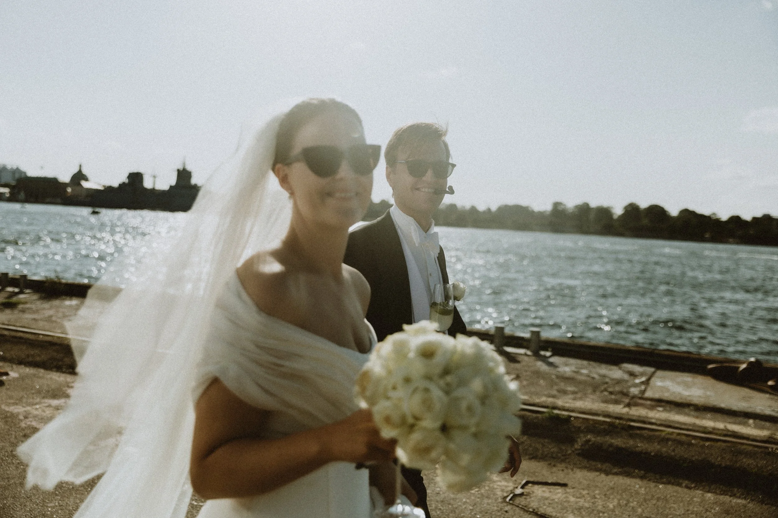 En nygiftepar på Københavns Havn, med vandet i baggrunden, begge iført solbriller, glad og smilende.