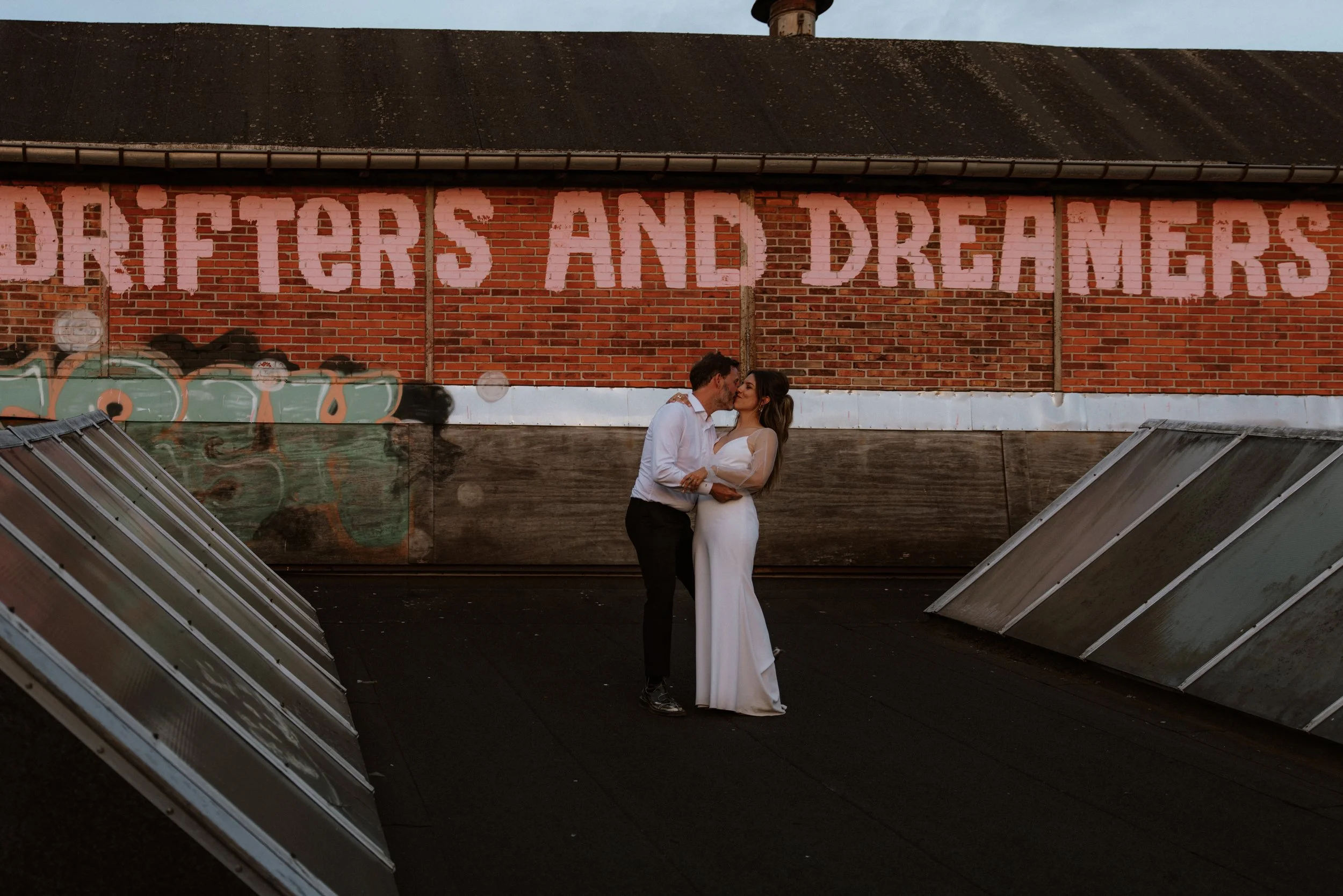 Et par i bryllupsoutfits står tæt sammen på en tagterrasse med solpaneler, foran en mur med graffiti og teksten 'DREAMERS AND DAREDEVILS'.