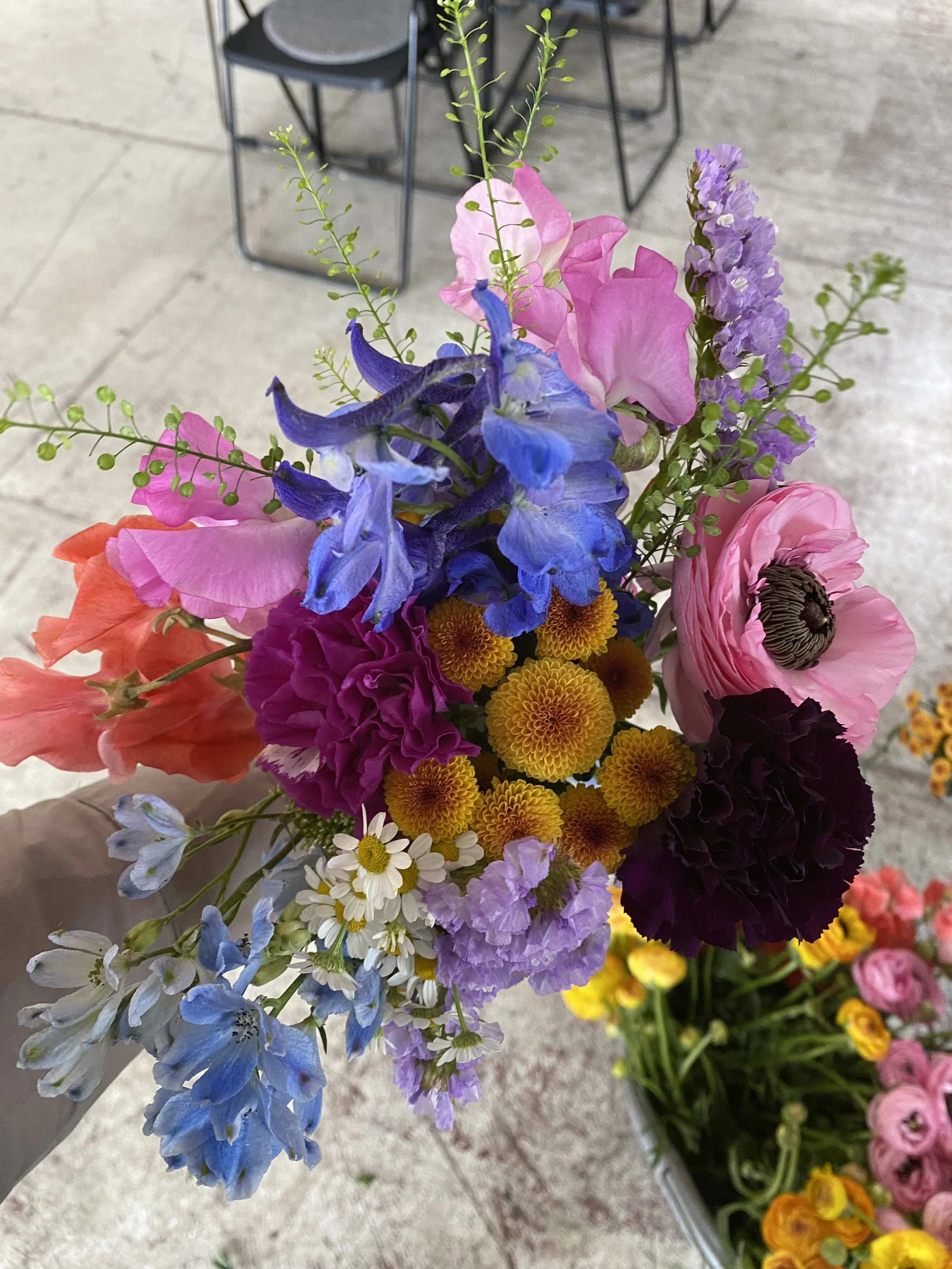 En buket fargerige blomster med blå, pink, lilla, orange, gul, hvid og mørk lilla blomster, taget på et indendørs sted.