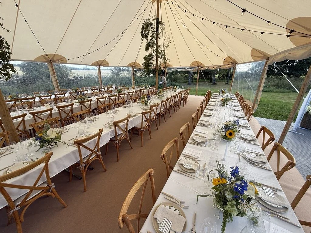 Bryllupsmiddag med langt bord dækket med hvide duge, blomsterarrangementer, tallerkener, For Holmegaard glas og Sabre Bistro Hornbestik under et luksustelt i haven.