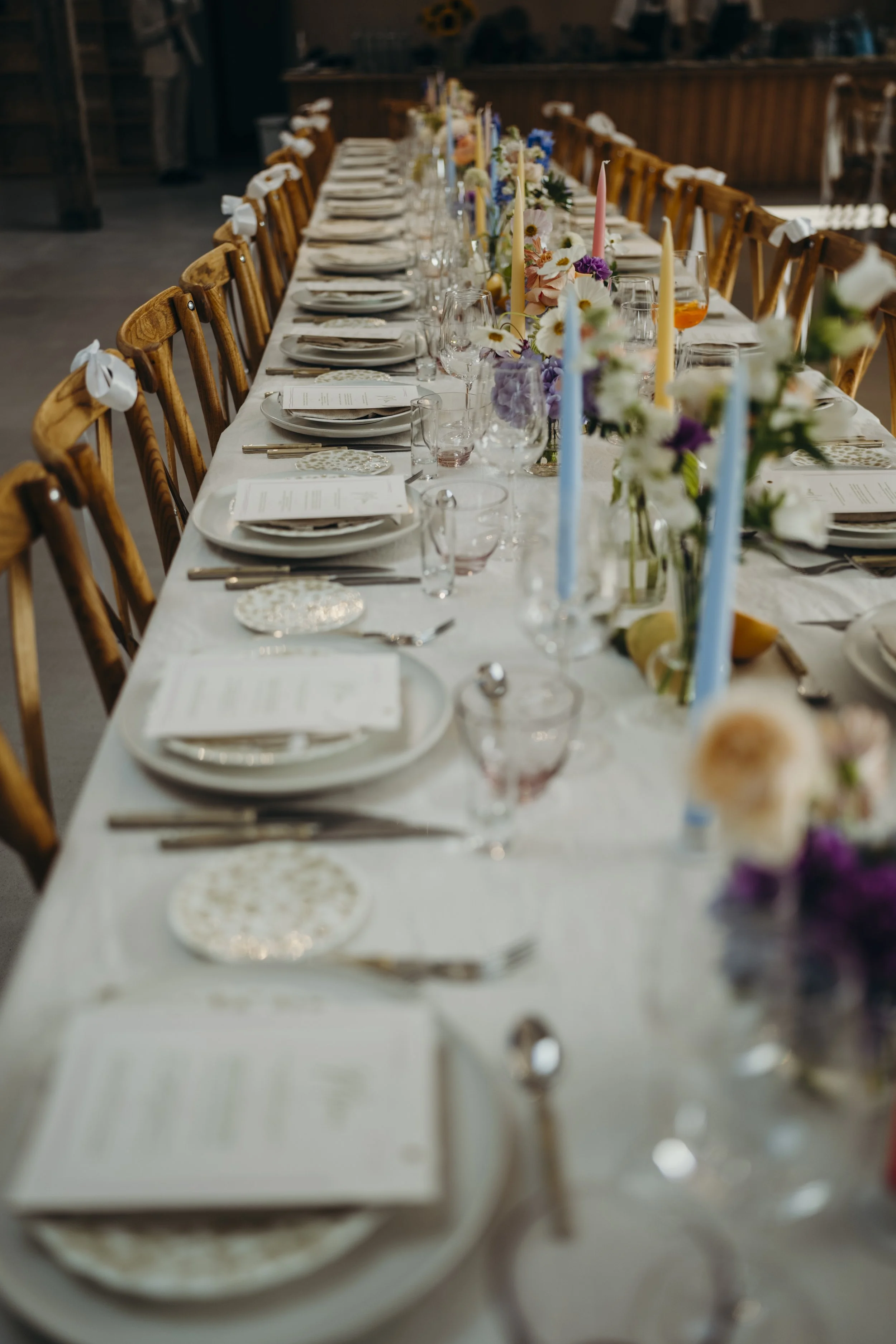 Lang bordret med spisebestik, tallerkener, glas, lyslåg, blomsterdekorationer og farvede stearinlys.