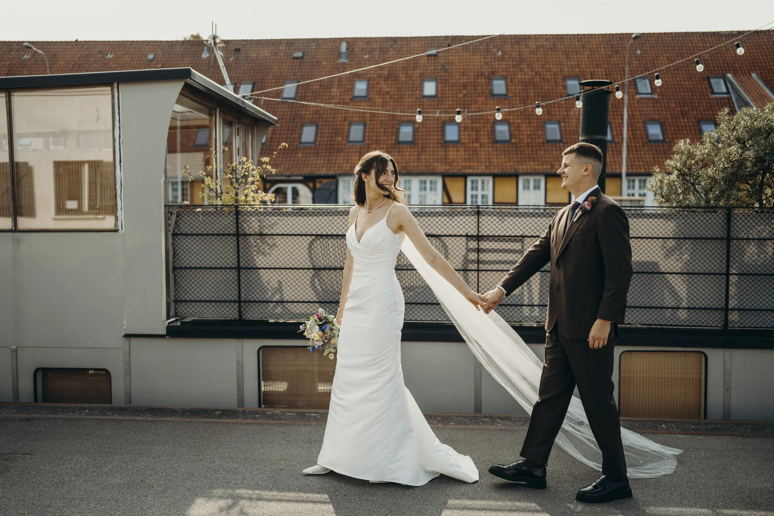 Et brudepar holder hænder på en udendørs sted, med en kvinde i en hvid brudekjole og en mand i en sort habit, solen skinner, og der er bolighuse i baggrunden.