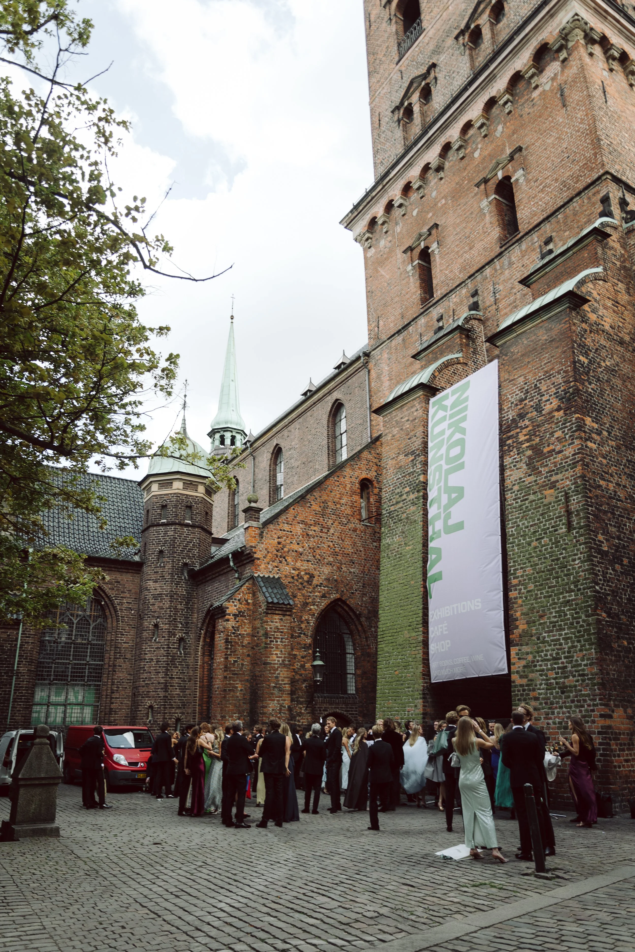 Folk i festtøj står udenfor Nikolaj Kunsthal, gammel, murstensbygning, det er et kunstgalleri. Der er en stor banner, der siger 'NORDIC ART EXHIBITION'.