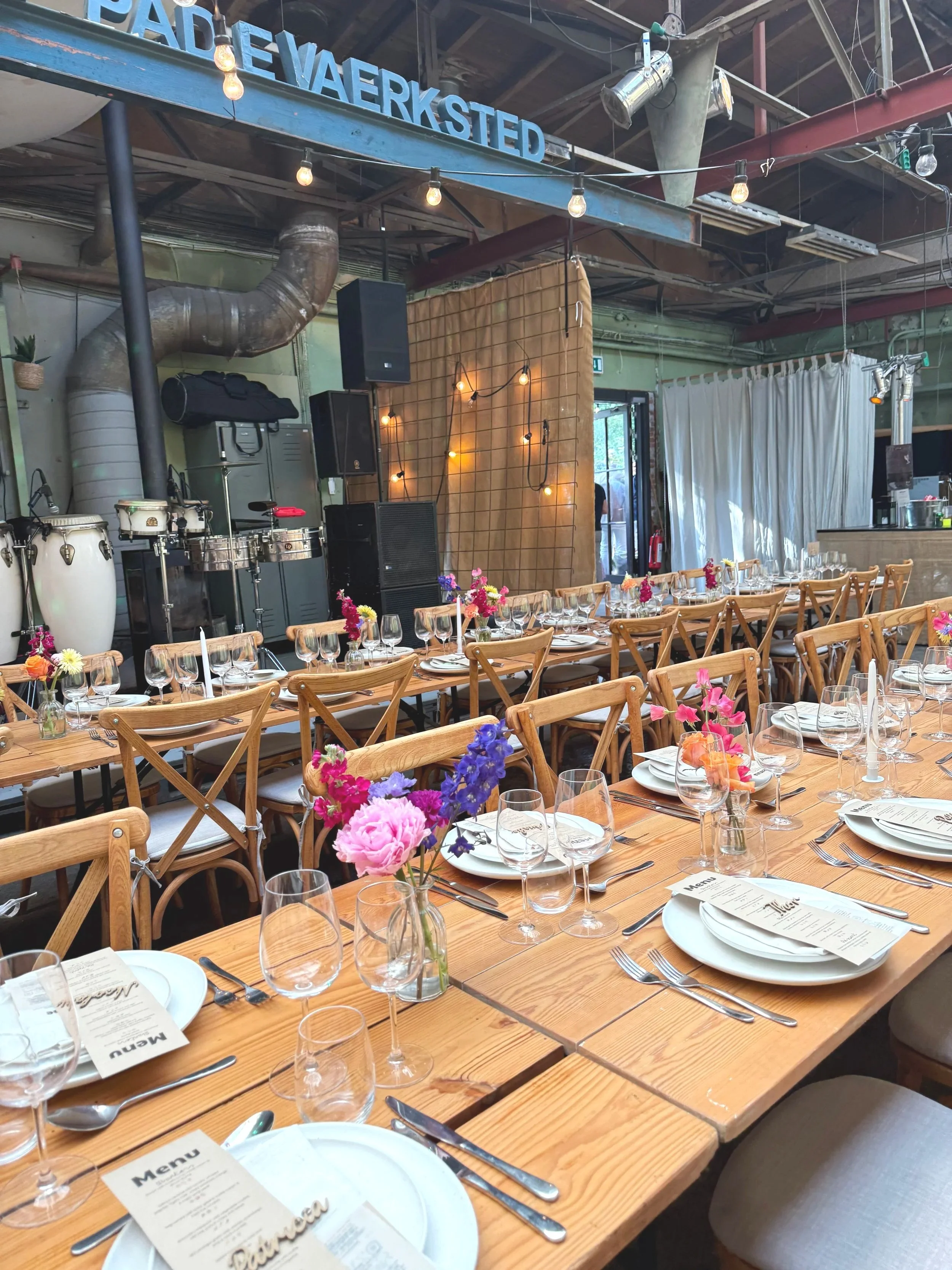 Indretning af et eventlokale med langborde dekoreret med blomster, glas og tallerkener. Der er en scene med musikinstrumenter og advarselsbelysning. En blå skilt med teksten 'PÆDE VAERKSTED' hænger over.