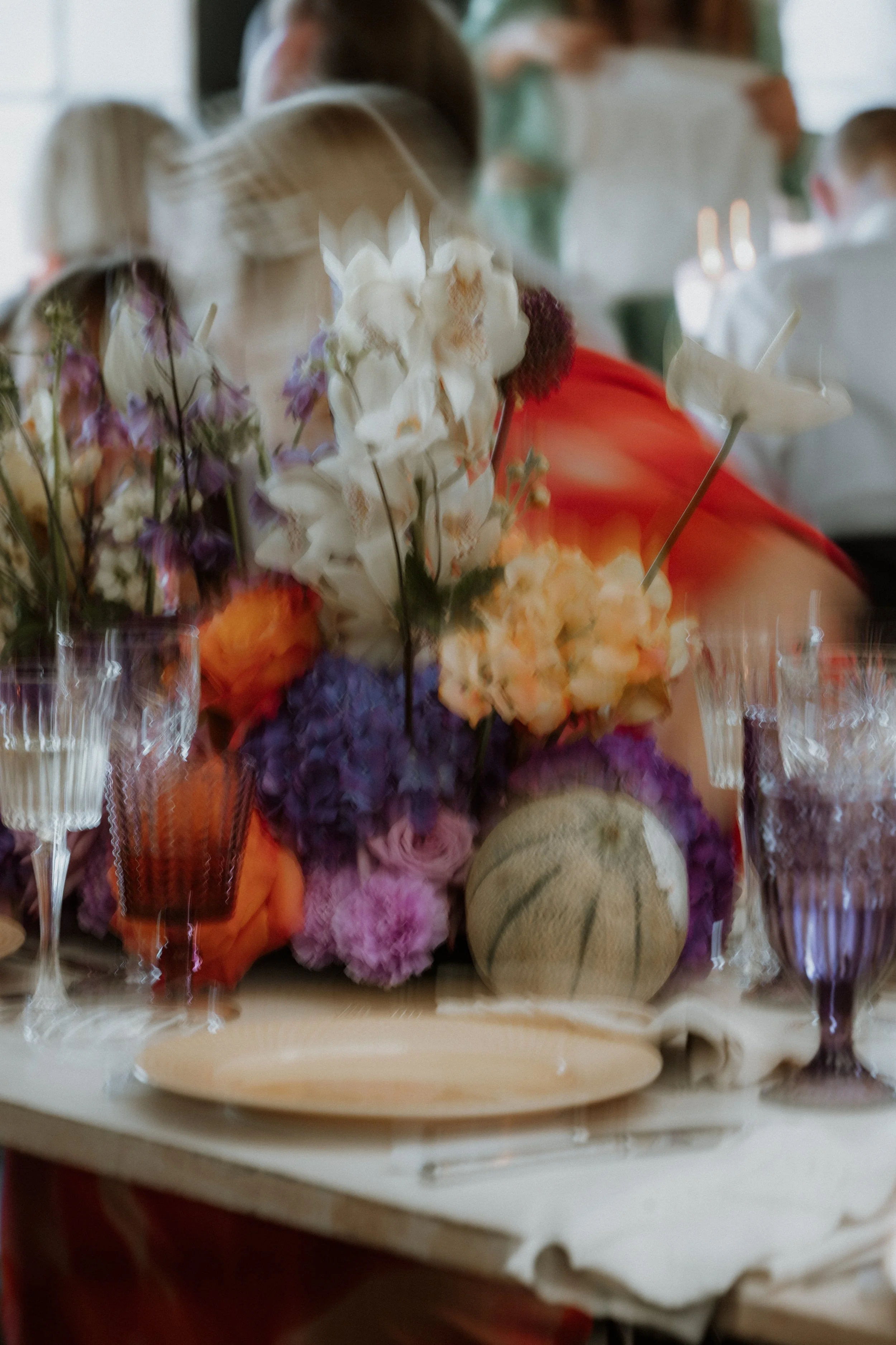 Billedet viser en borddækning med en stor blomsterdekoration bestående af forskellige blomster i lilla, hvid og orange nuancer. Der er også et melon på bordet. Baggrunden viser folk, der sidder ved bordet, nogle med korte blond hår.