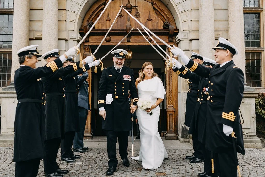 En bryllupspar går gennem en militærparade udenfor en historisk bygning, omgivet af uniformklædte militærpersoner, der holder sabler hevet op i luften.