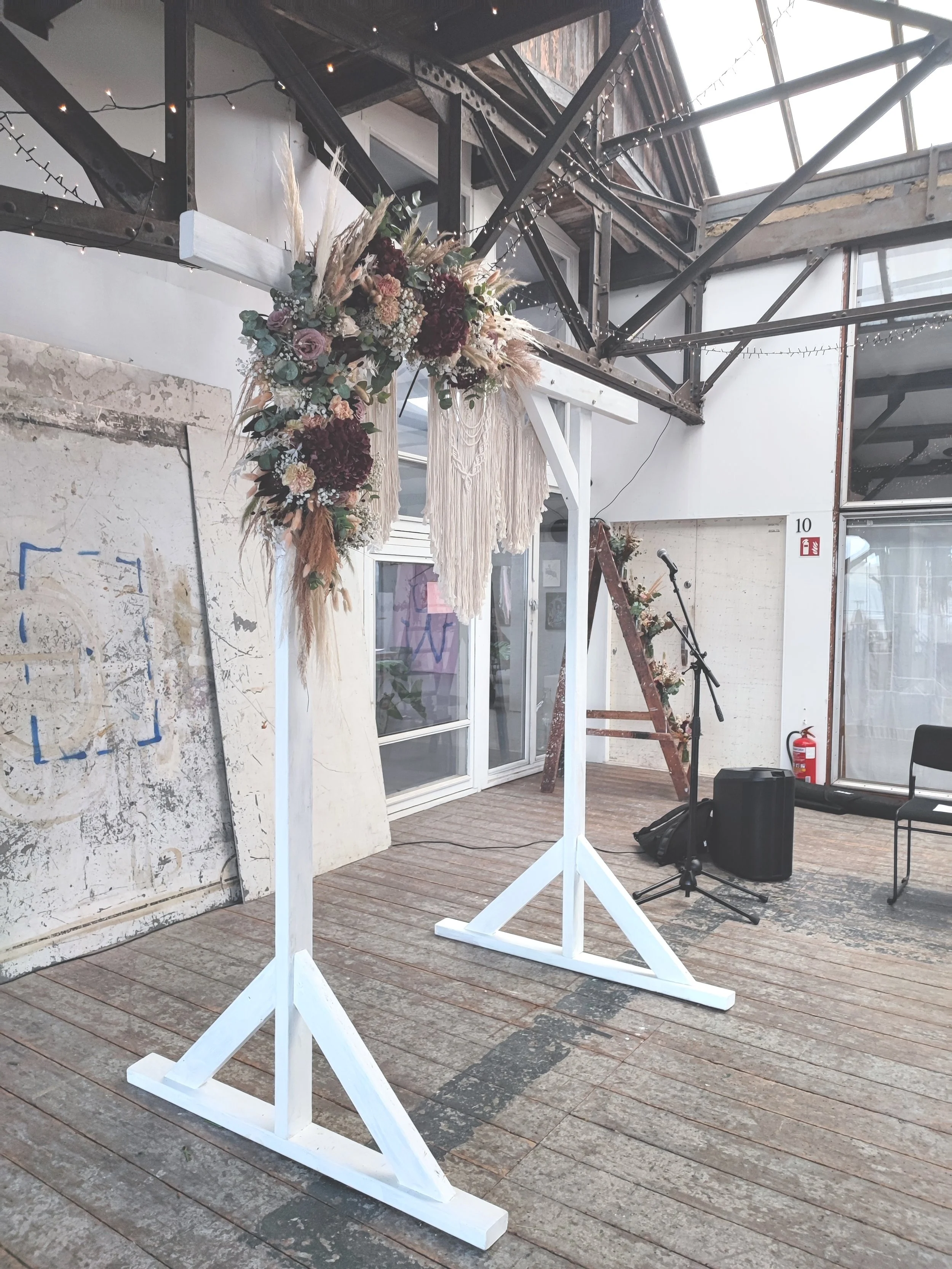 Blomsterdekoration på æstetisk hjortestativ, klar til en ceremoni, med mikrofon og lydudstyr i baggrunden, inde i en industriel bygning.