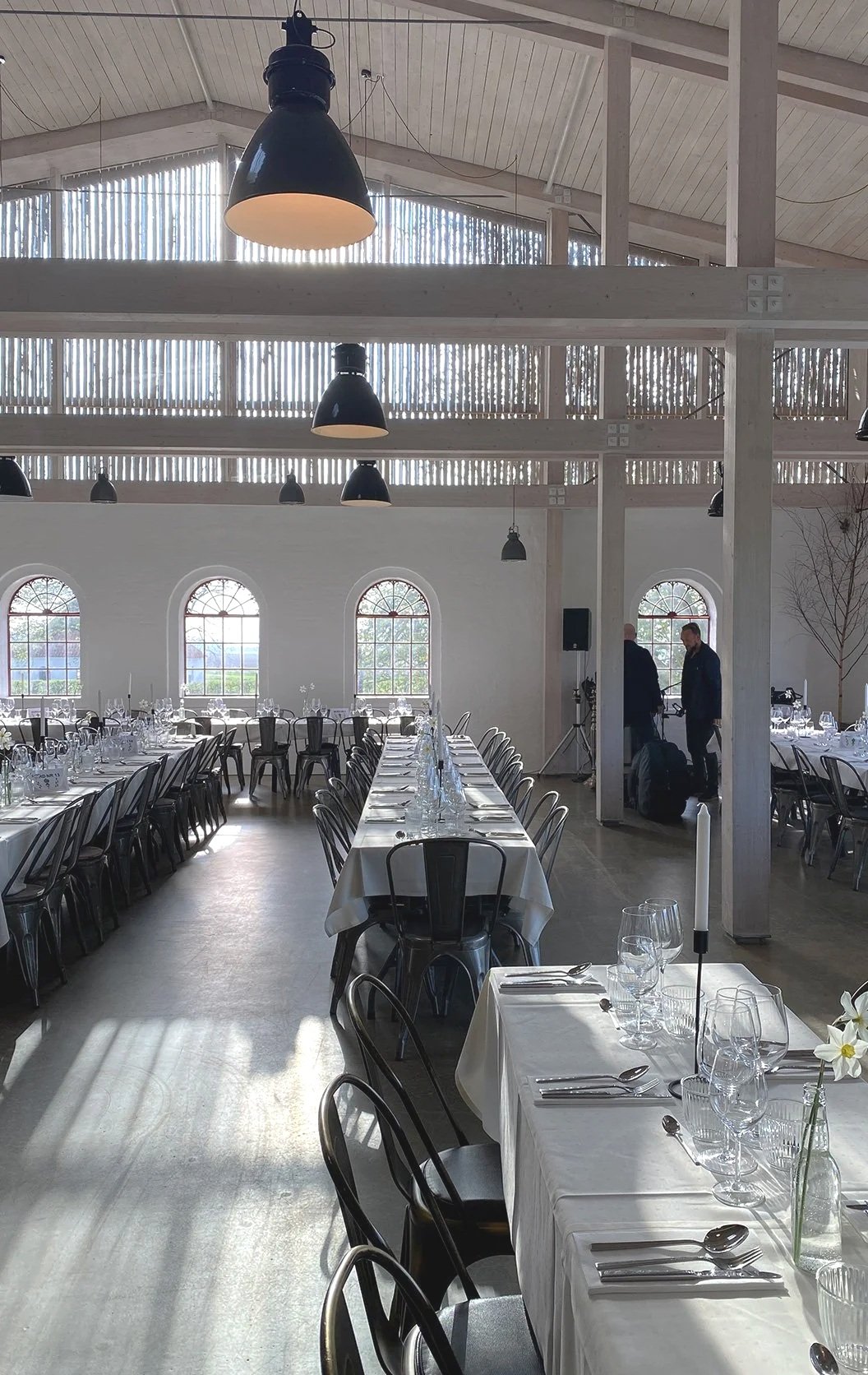 Søllestedgård eventloakle med langborde dækket med hvide duge, glas, bestik og blomster. Store vinduer og sorte lamper hænger fra loftet.