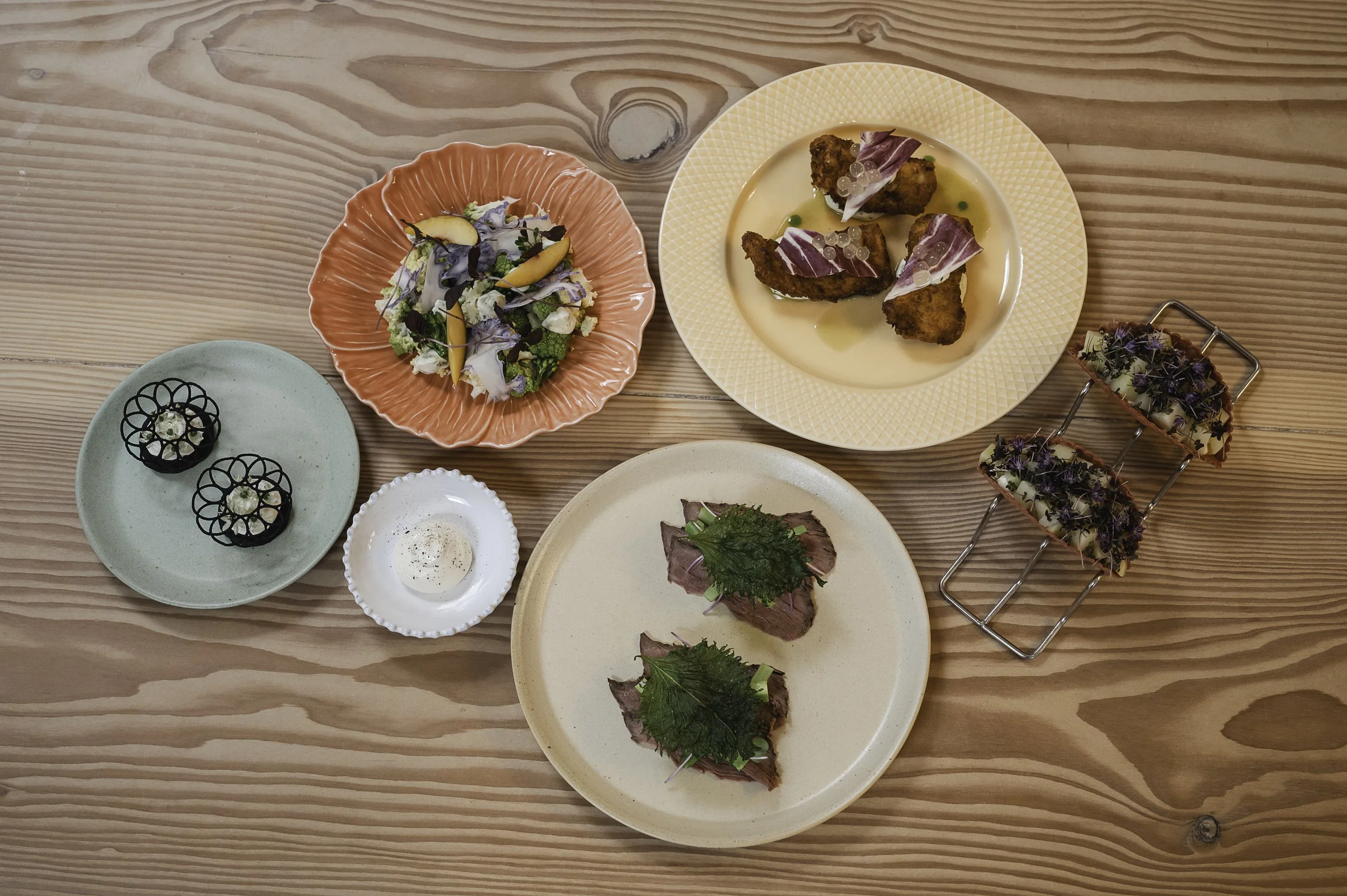 Billedet viser en række små retter på forskellige tallerkener, herunder en salat, to skiver oksekød med sauce, tre små retter med blomster, og nogle friturestegte kødgenstande med skinke på toppen, alle arrangeret på et træbord.