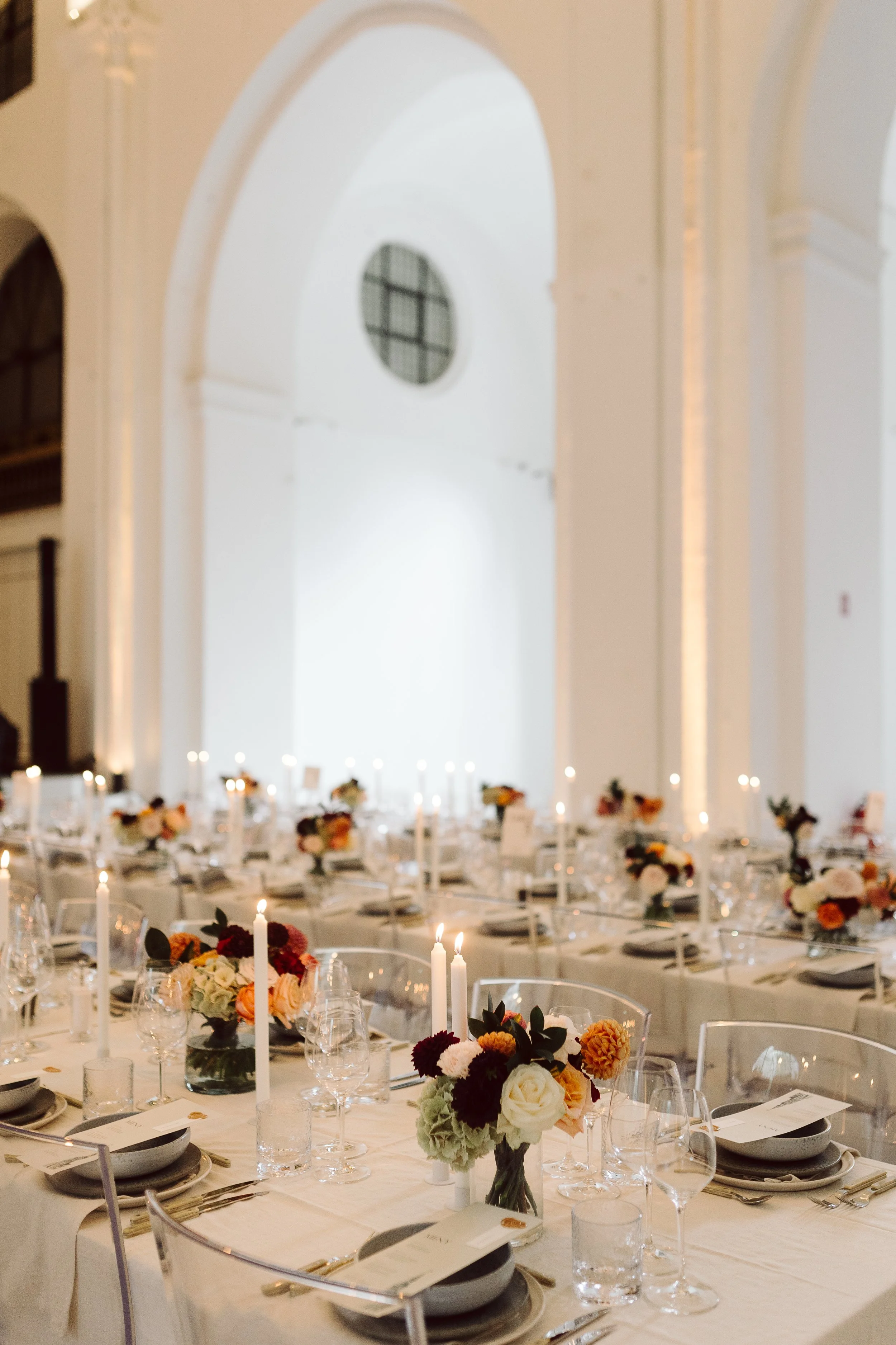 Elegant middagsbord med blomsterdekorationer og stearinlys i en stor, lys sal med hvide vægge og store arkader.