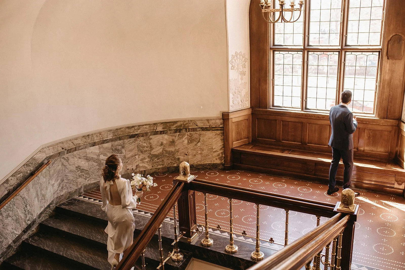 En kvinde med mavebælte holder en blomsterbuket, mens en mand i habit stående ved et stort vindue ser ud. Stedet er elegant indrettet med træpaneler, en stor vinduesramme, og en trappe med guldende gelænder.