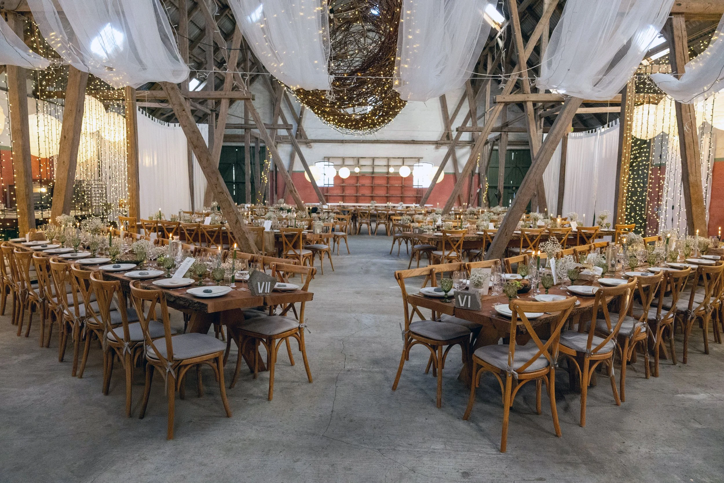 Indendørs festlokale med rundborde, dekoreret med blomster, lys og servietter, omgivet af trælade med hvide gardiner og lysbelysning.