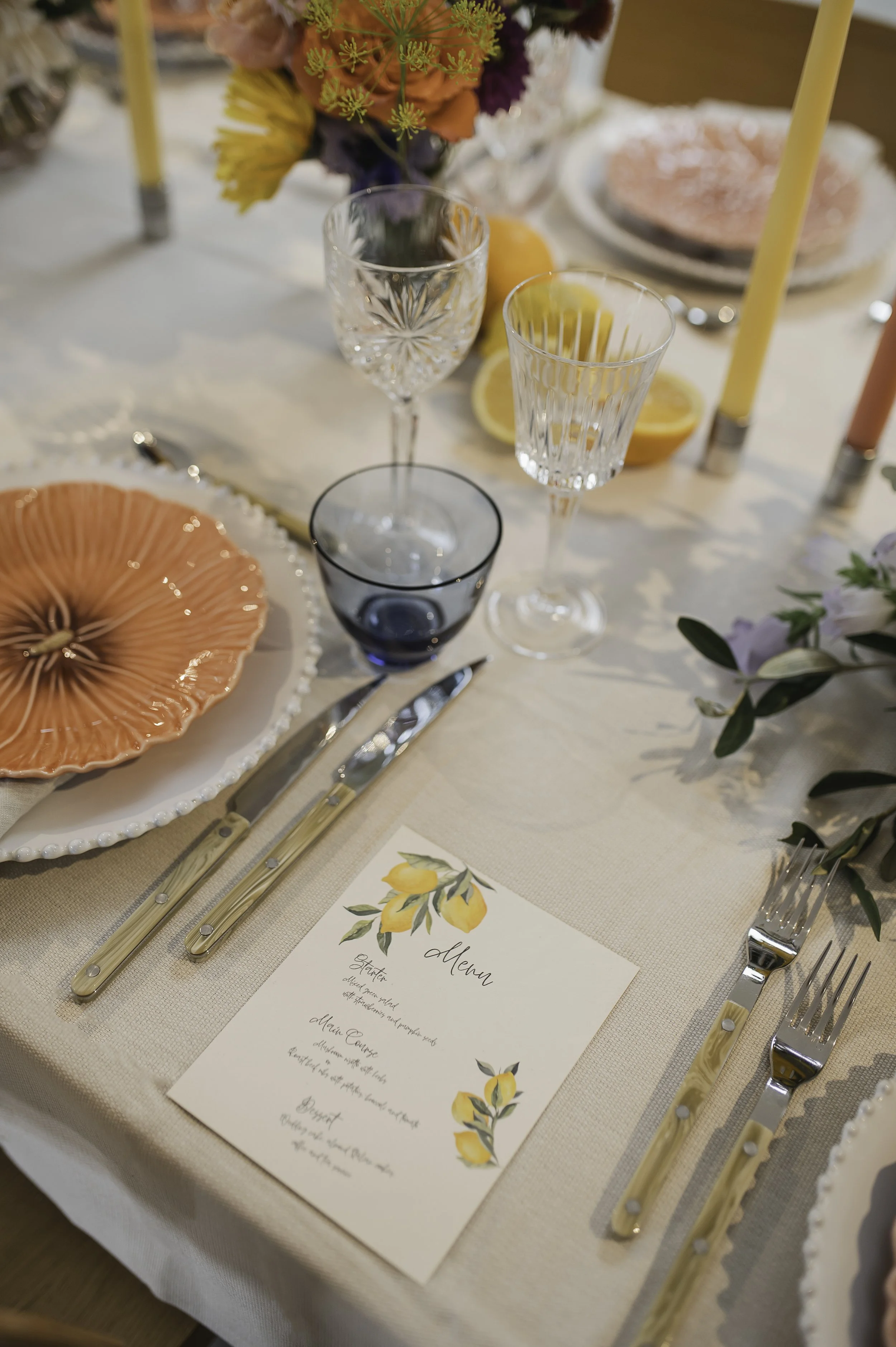 Elegant borddækning af Bliss Cph med Bordallo Pinheiro middagstallerkener, Sabre Bistro Horn bestik, vin- og vandglas, en menu med citroner, blomsterdekorationer og tændte stearinlys.