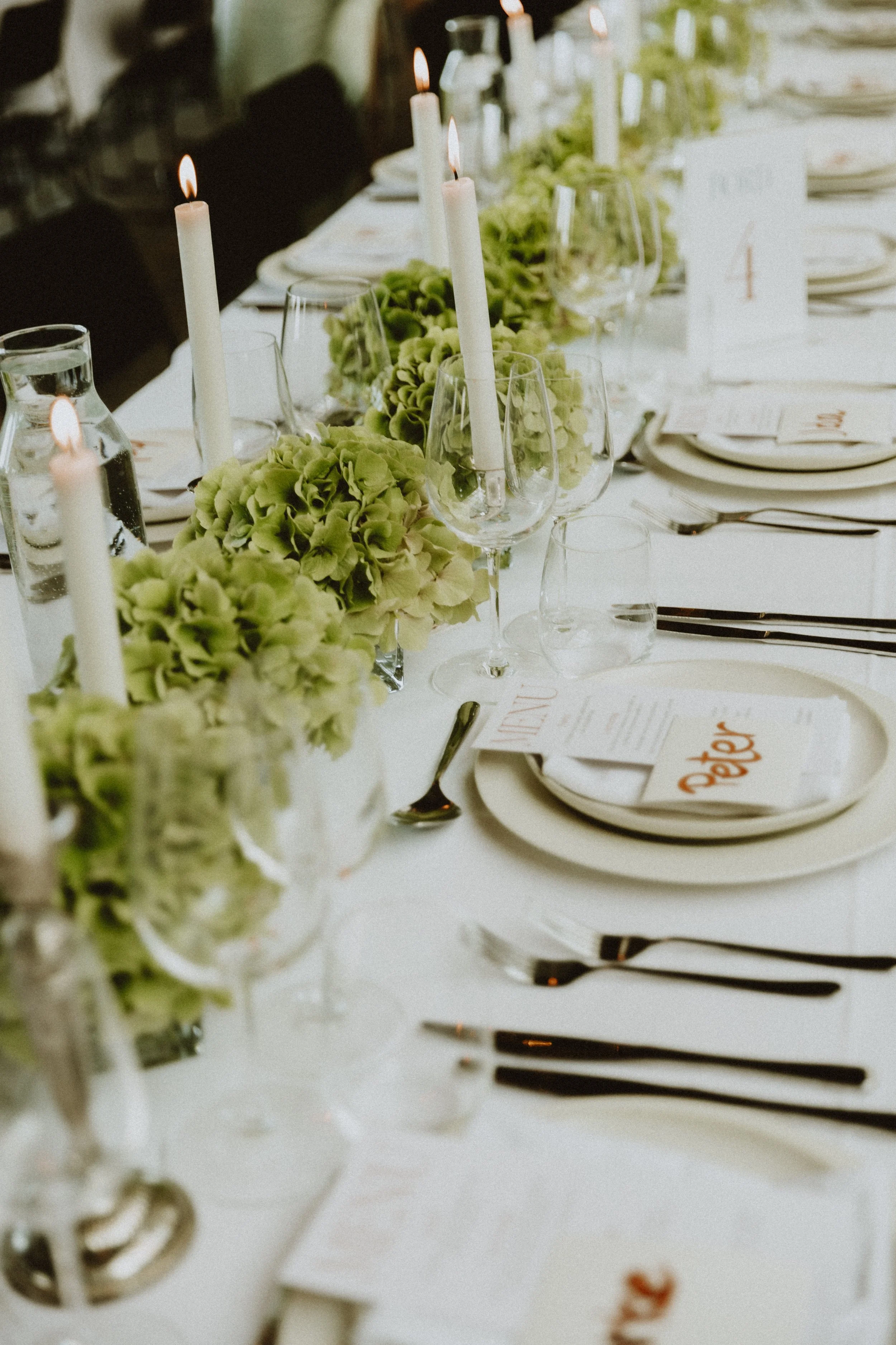 Elegant festborddekoration med grønne blomster, hvide stearinlys og elegante glas og tallerkener.