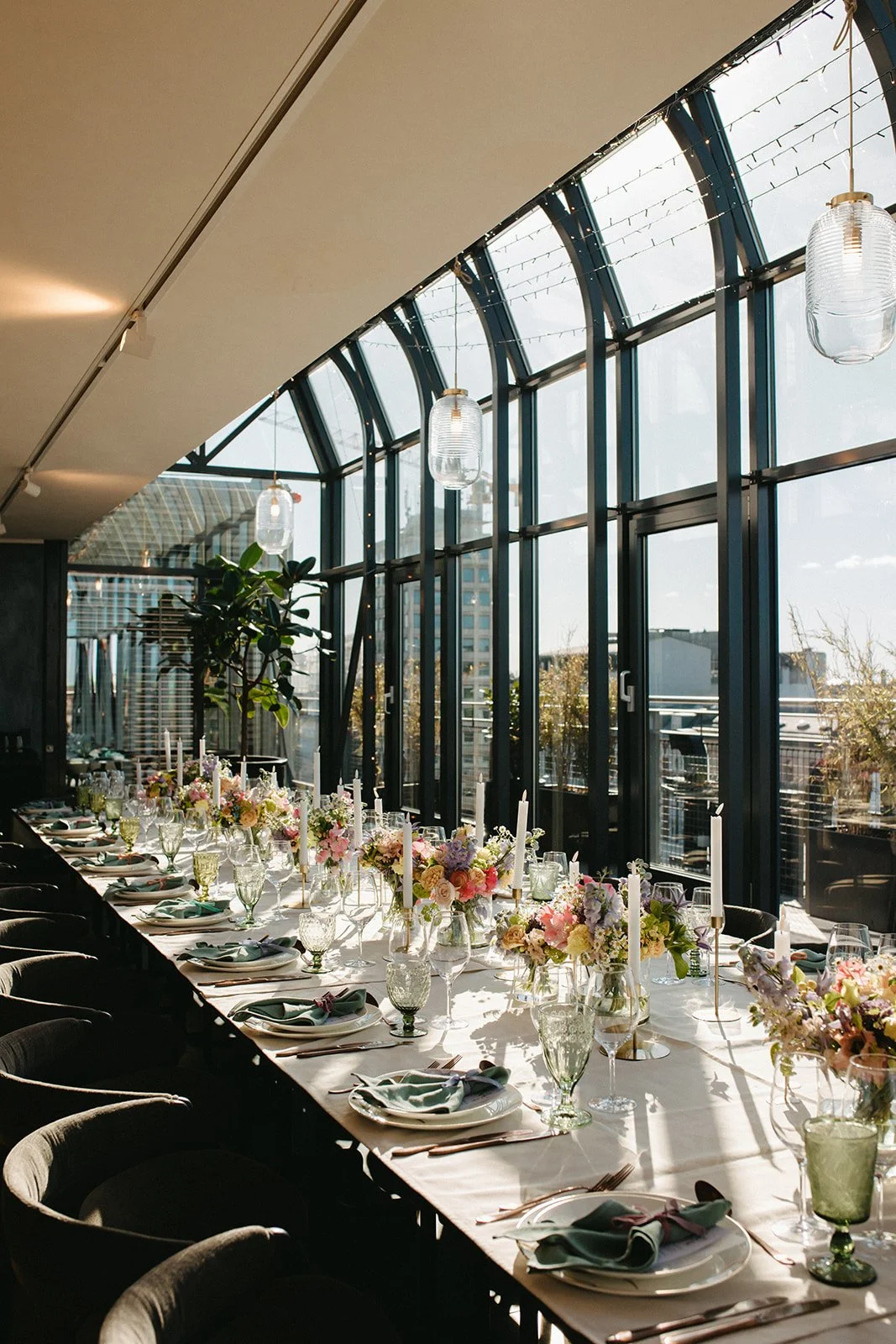 Bliss CPH – På Djøf Rooftop en lang borddækning med blomsterdekorationer, lysestager, tallerkener, glas og bestik i et lyst rum med store vindue