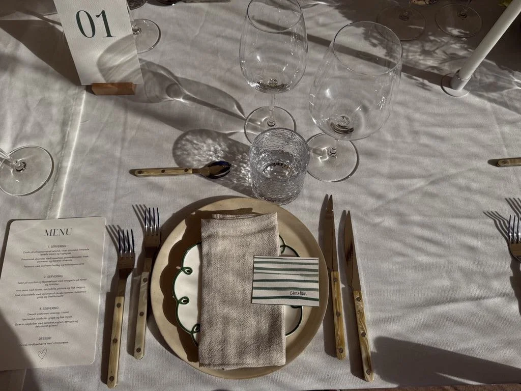 Borddækning med tallerken, bestik, tynde klare glas, håndmalede navneskilte og menu, på en hvid dug, med skygger fra sollys