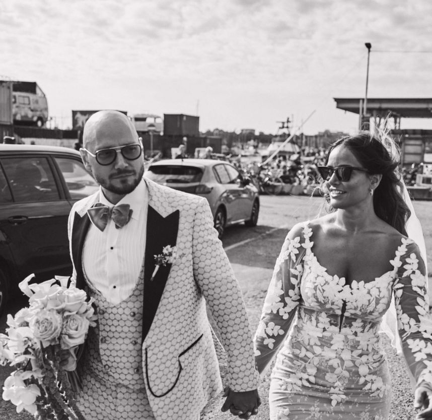 Sort-hvid foto af et bryllup med en mand i en hvid og sort jakkesæt med blomster på brystlommen og en bære af blomster, og en kvinde i en blomsterdetaljeret kjole og solbriller, der går hånd i hånd udenfor.
