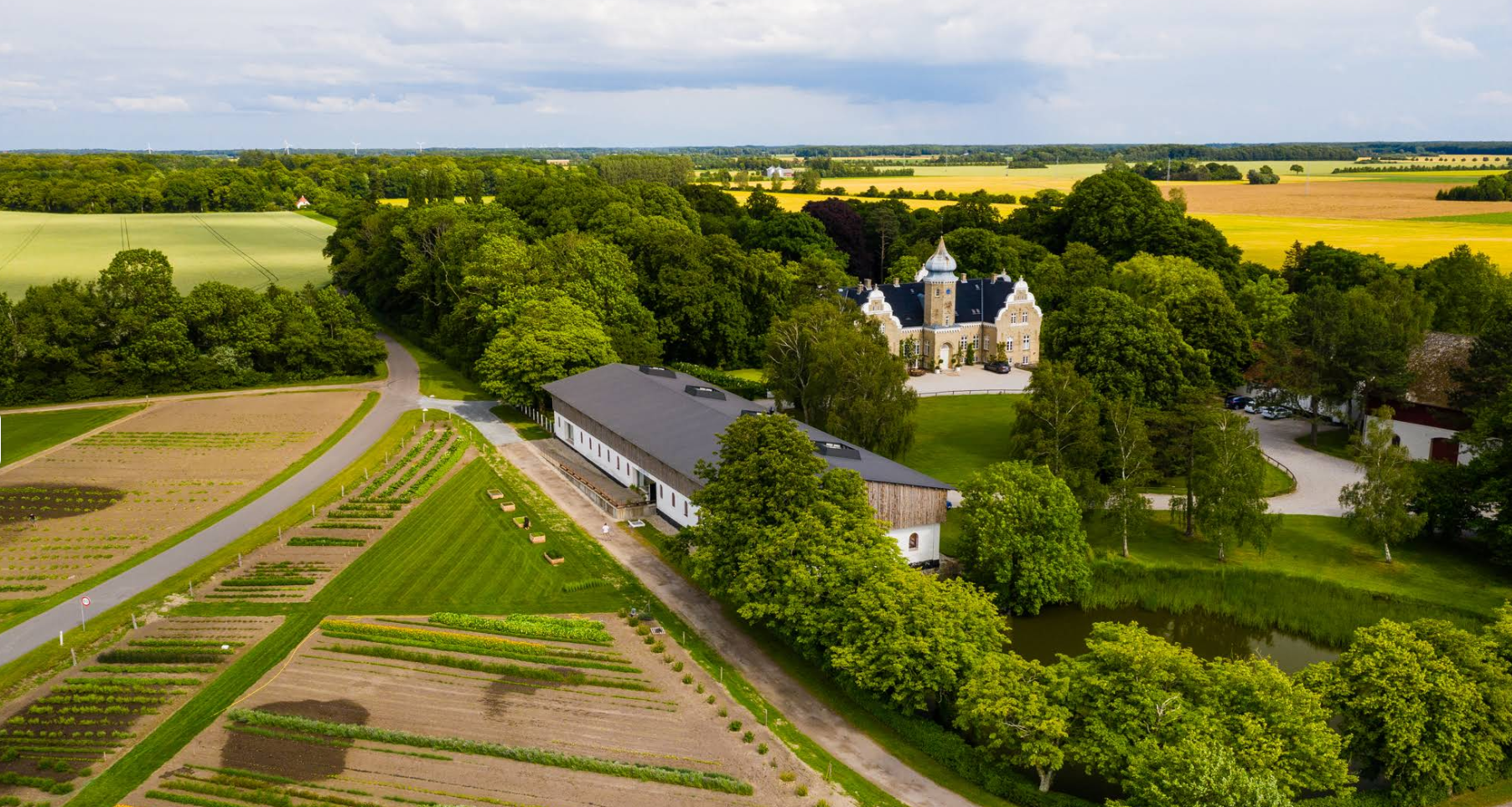 Bliss Cph - Grøn landlig ejendom, Søllestedgård set ovenfra, et langt hvidt bryllupssted, grønne marker, skov og en lille sø.